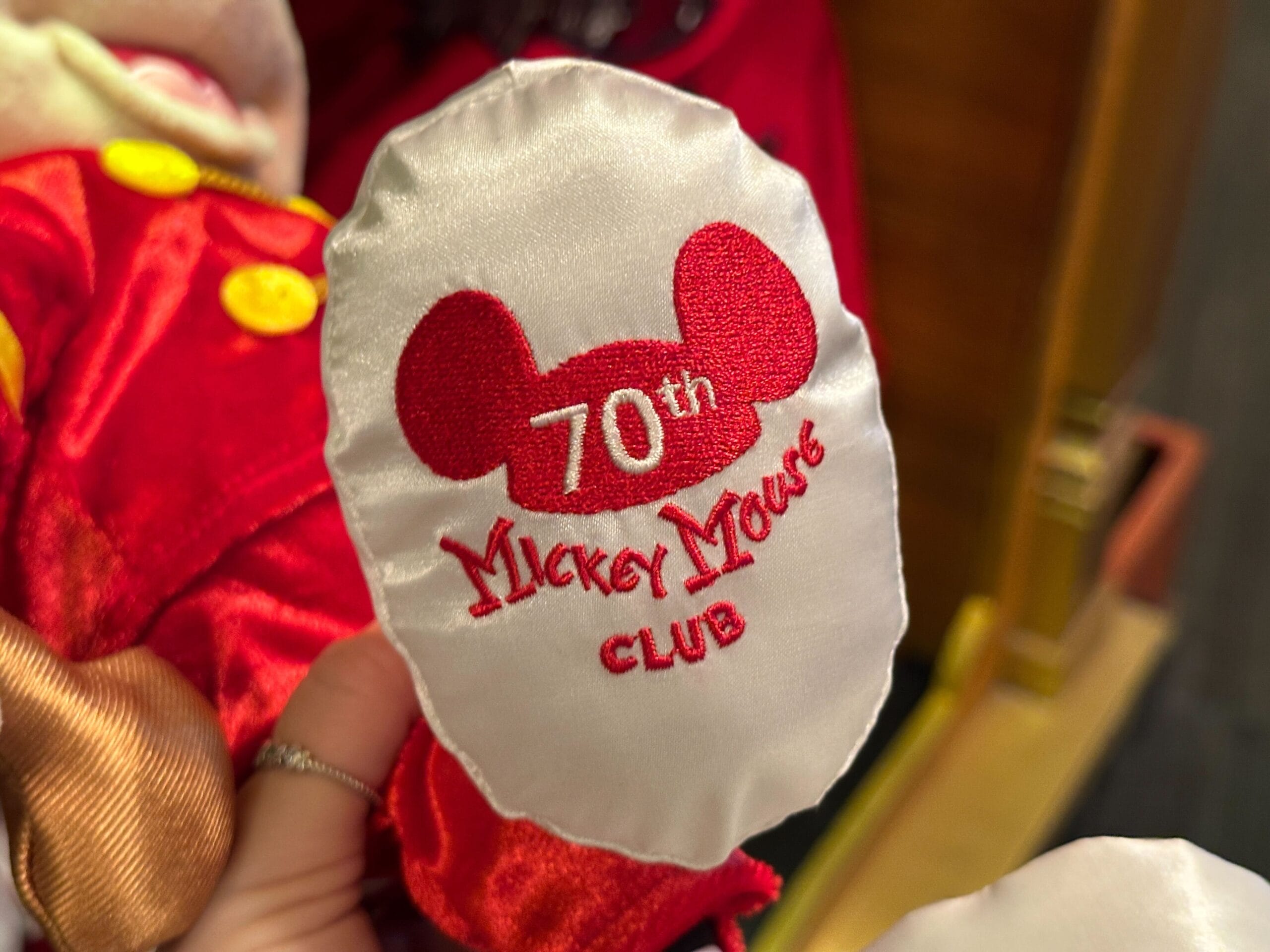 White satin patch with red Mickey ears and text: "70th Mickey Mouse Club" held by a hand.