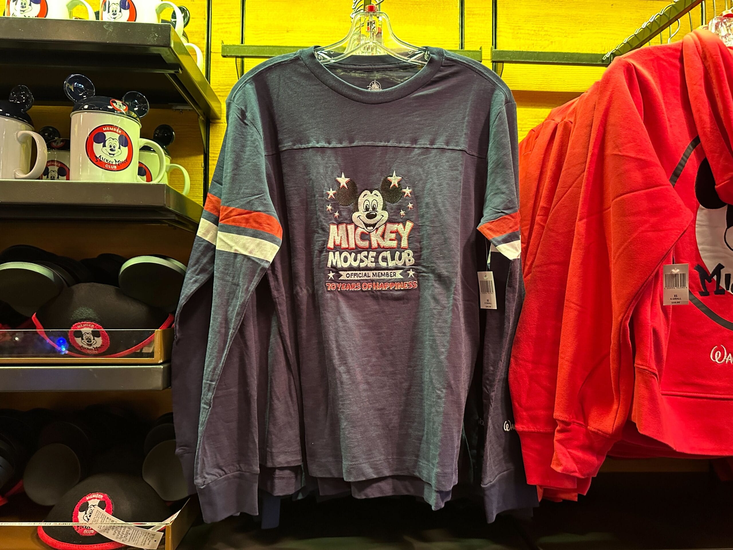 Navy blue Mickey Mouse Club sweatshirt with stripes on sleeves displayed on a rack in a Disney store.
