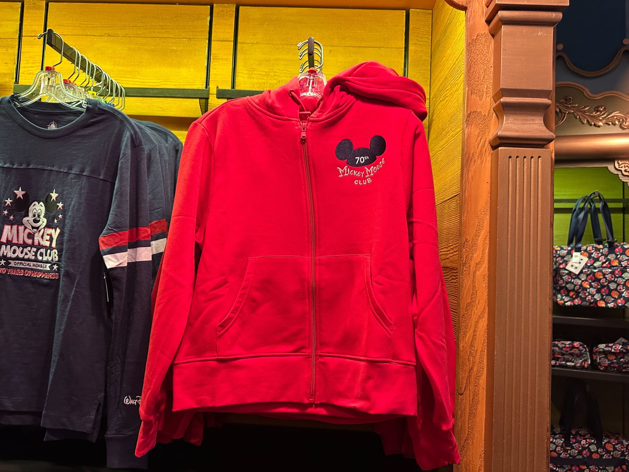 A red zip-up hoodie with a Mickey Mouse Club logo hangs on a rack in a store next to other Disney apparel.