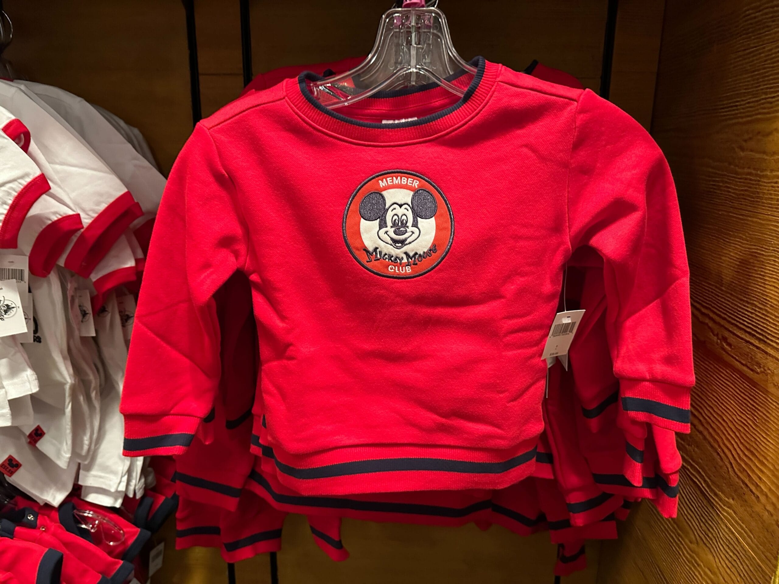 Red long-sleeve children's shirt with Mickey Mouse "Member" patch, hanging on a rack in a store.