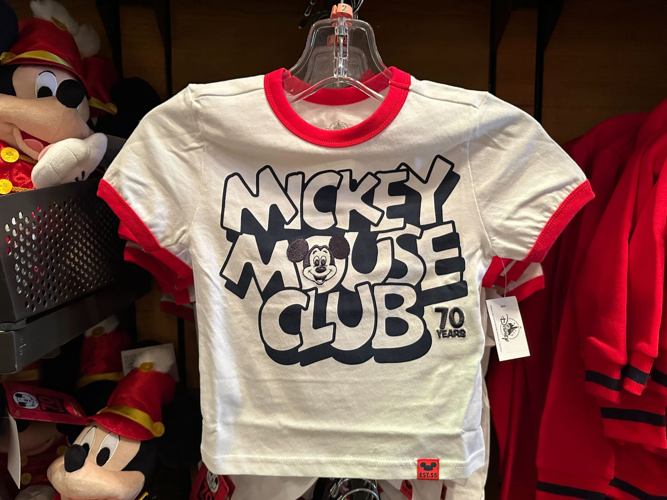 White t-shirt with red trim that says “Mickey Mouse Club 70 Years” in bold black letters, displayed on a hanger.