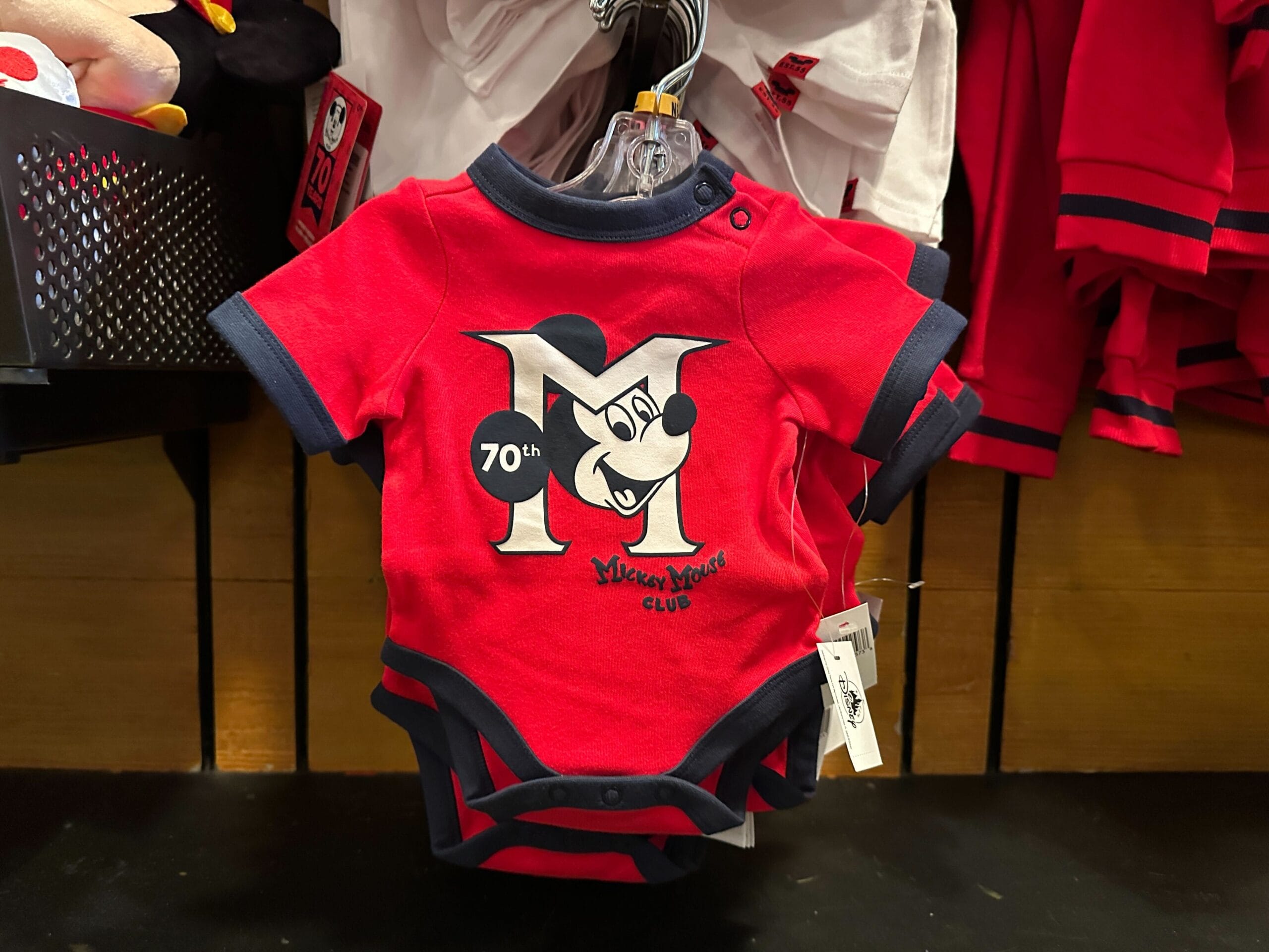 Red baby onesie with Mickey Mouse and "70th M Mickey Mouse Club" logo, hanging on a store display.