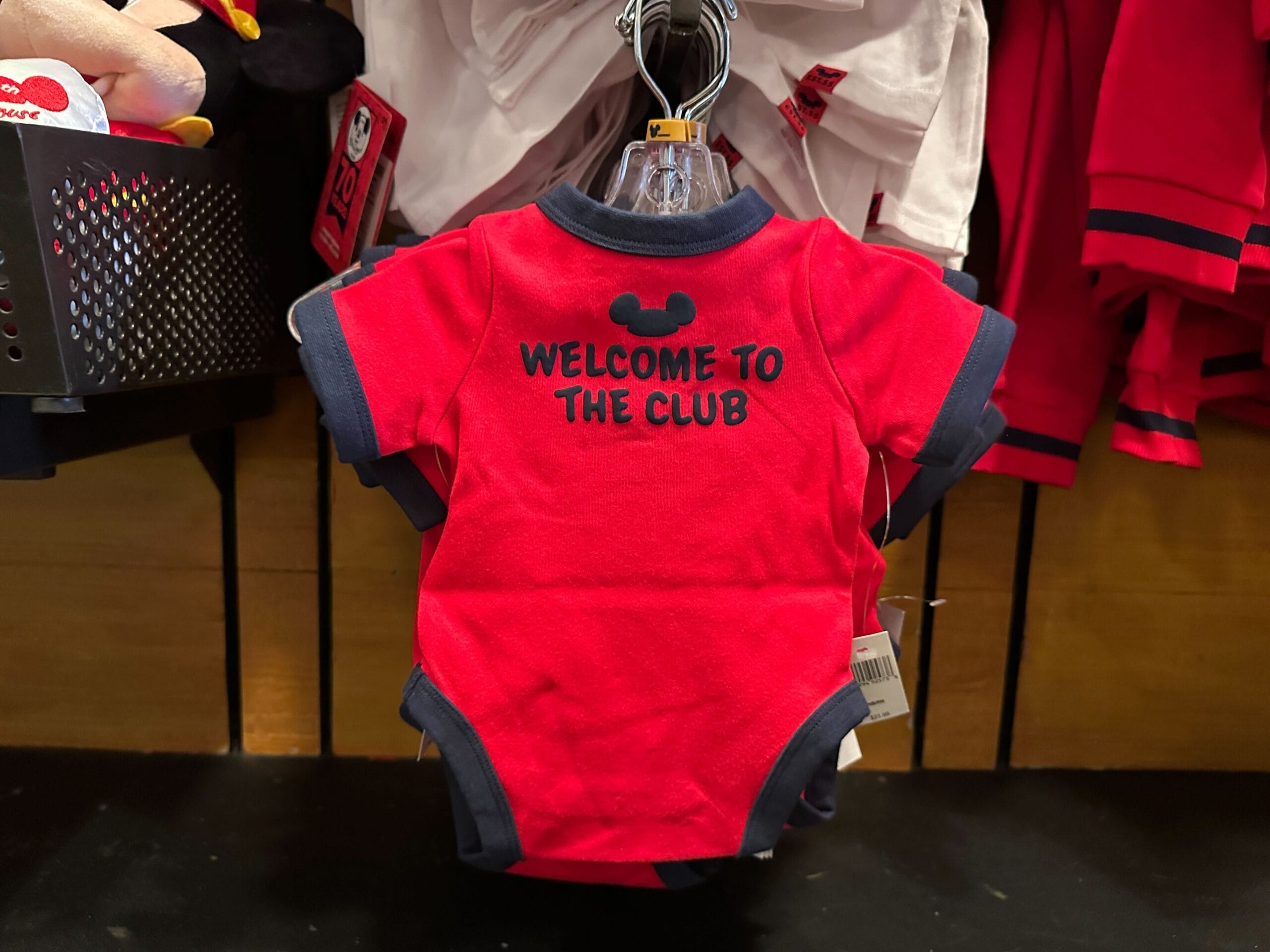 Red baby onesie with navy trim and “WELCOME TO THE CLUB” text, hanging on a display rack in a store.