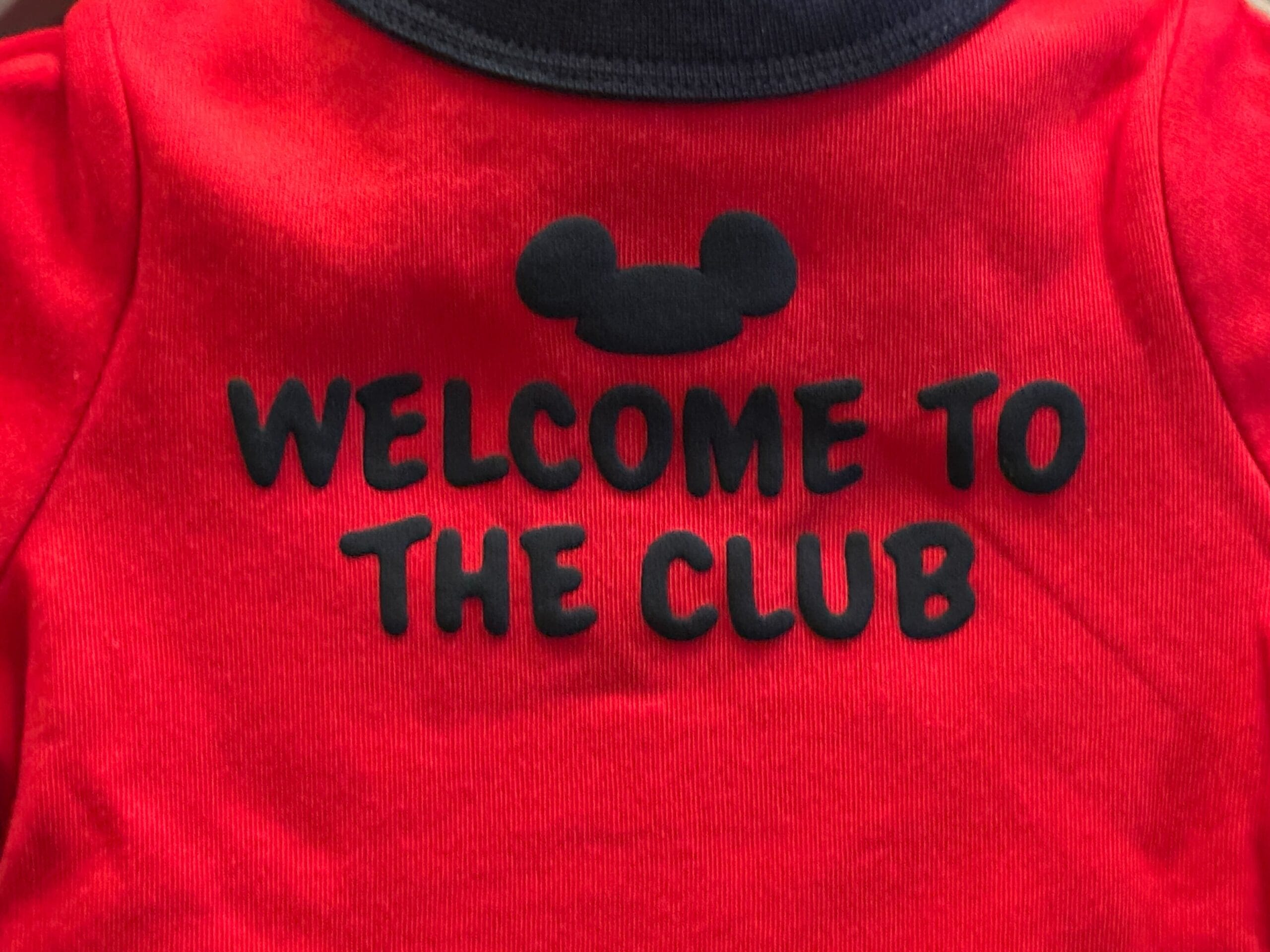 Red shirt with a black mouse ear silhouette and the text "WELCOME TO THE CLUB" in bold black letters.