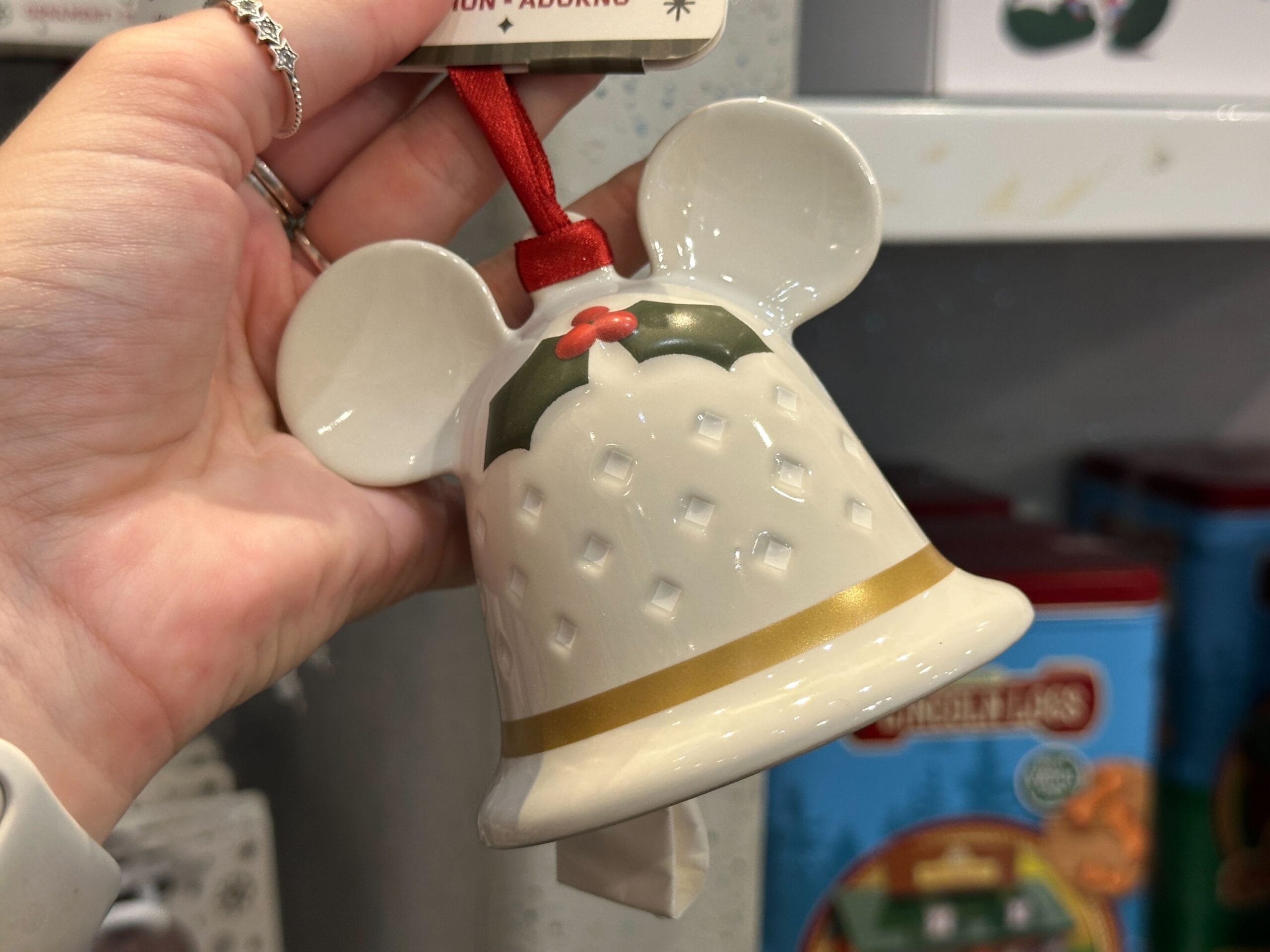 Hand holding a white ceramic bell Christmas ornament shaped like Mickey Mouse ears with gold and holly details.