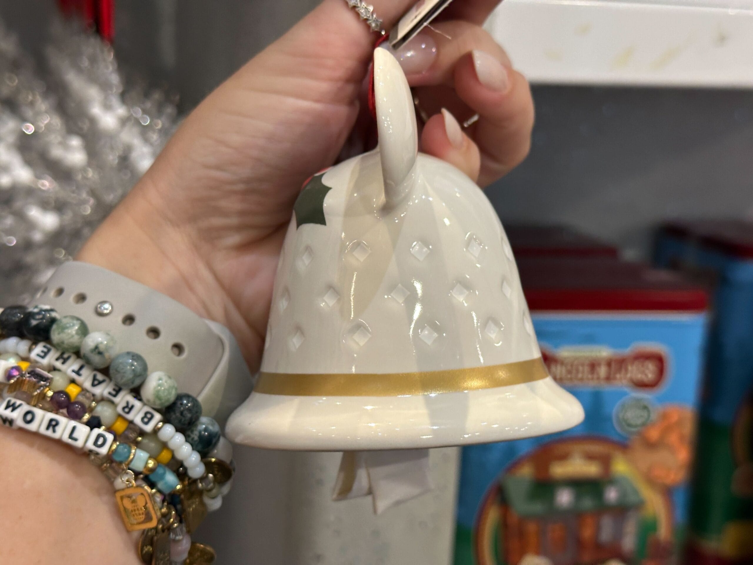 A hand holds a white ceramic bell ornament with gold trim and a diamond pattern, in front of holiday decorations.