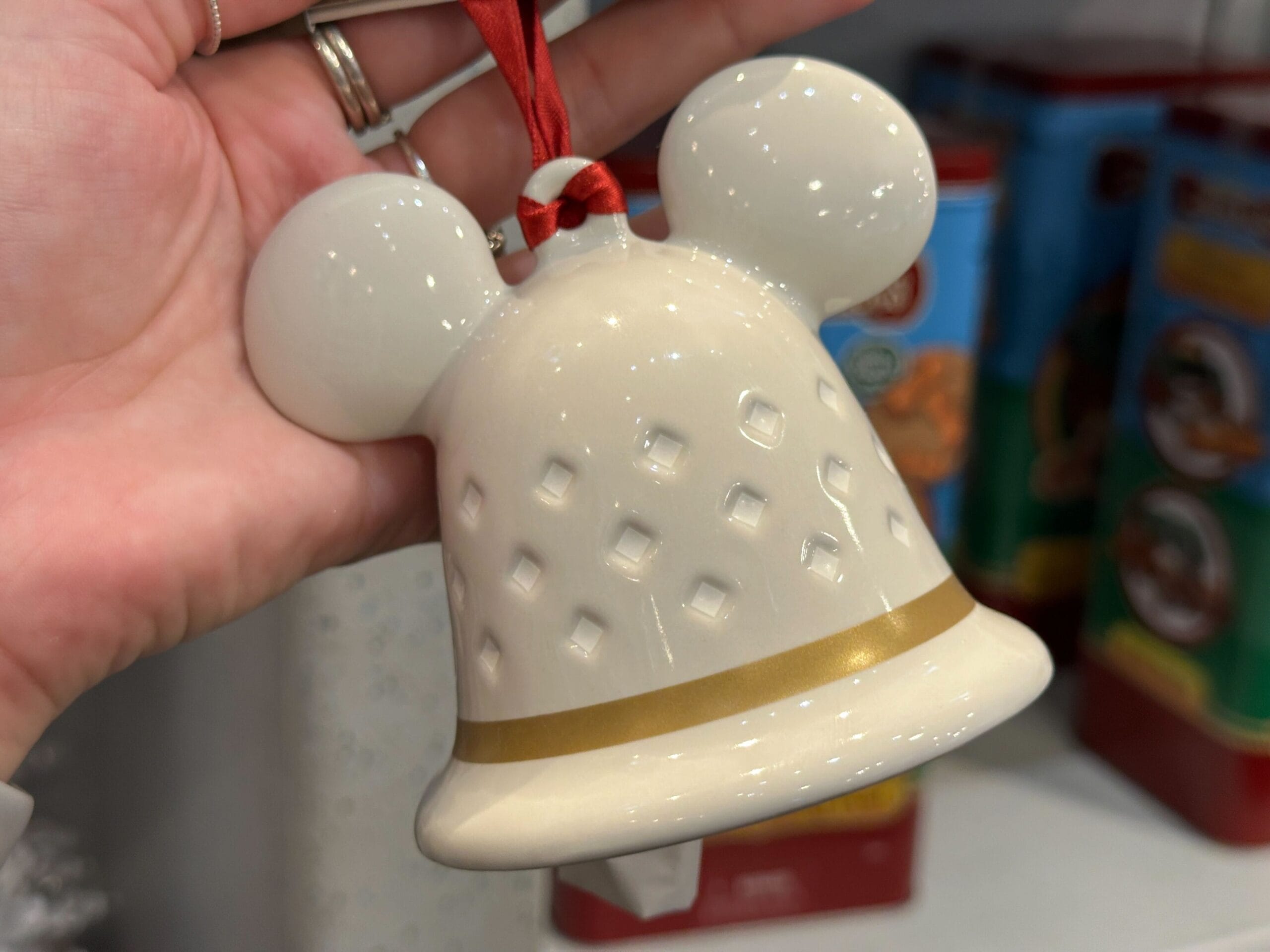 Hand holding a white ceramic bell ornament with mouse ears, gold stripe, and red ribbon; store shelf in background.
