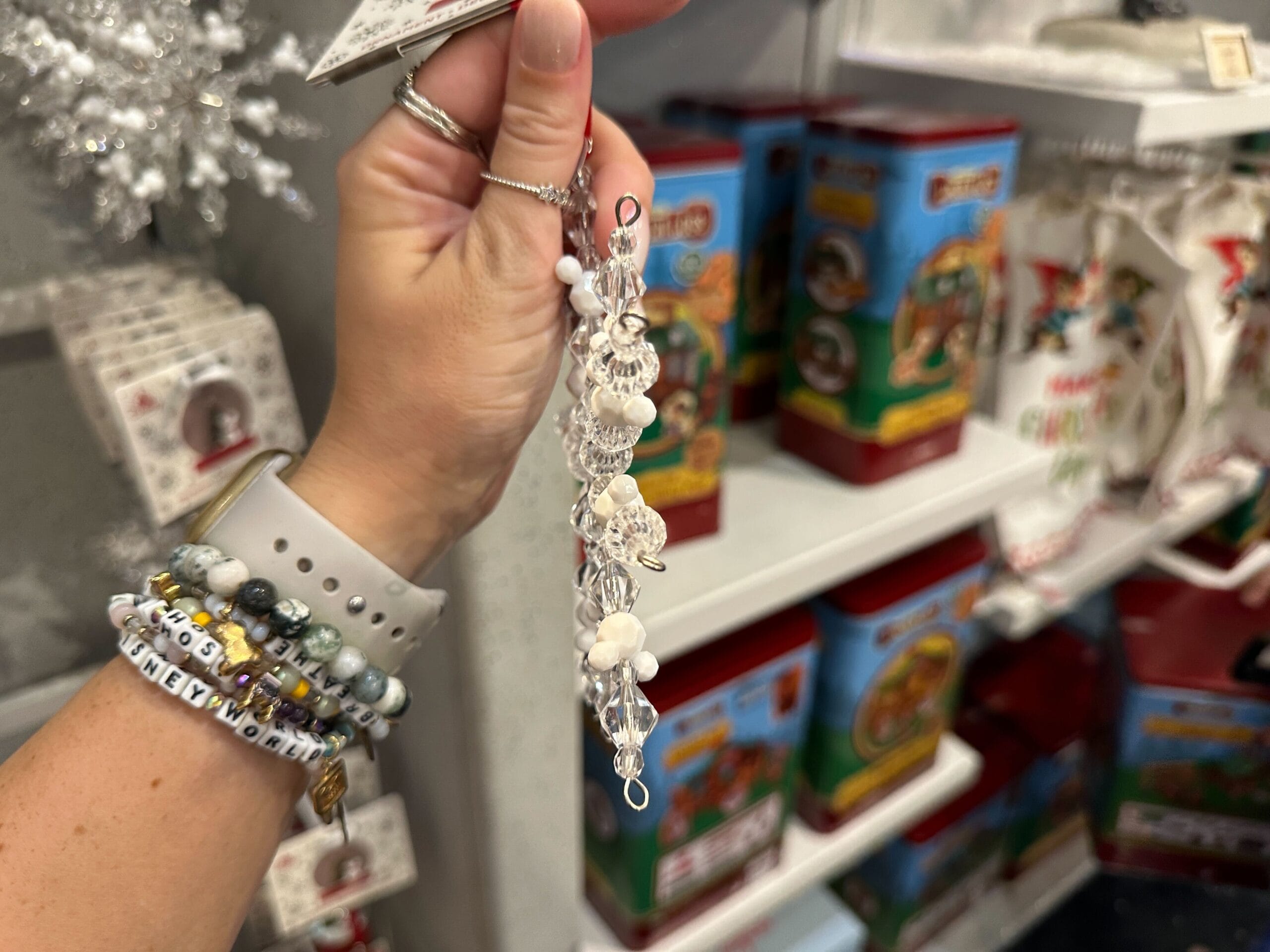 A hand wearing beaded bracelets holds a delicate beaded ornament in a store with holiday-themed items in the background.