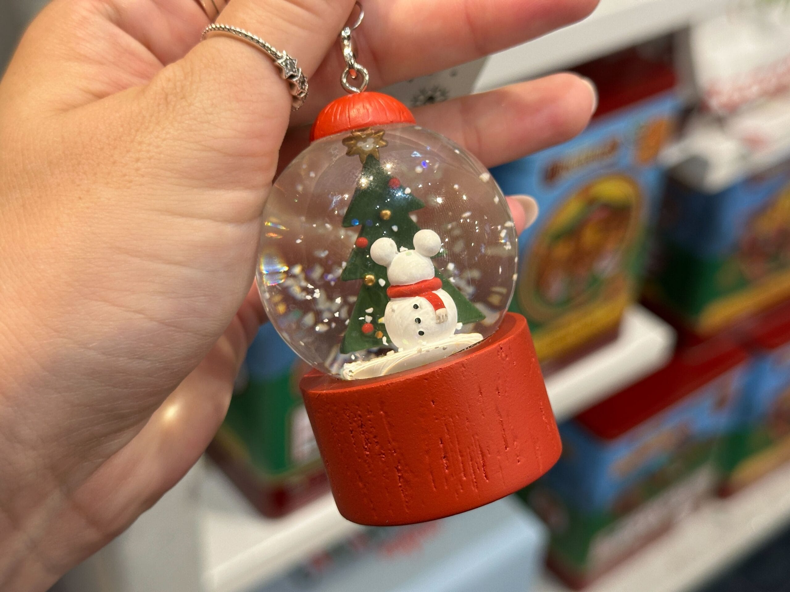 Hand holding a snow globe keychain with a snowman and Christmas tree inside, red base, and festive background.