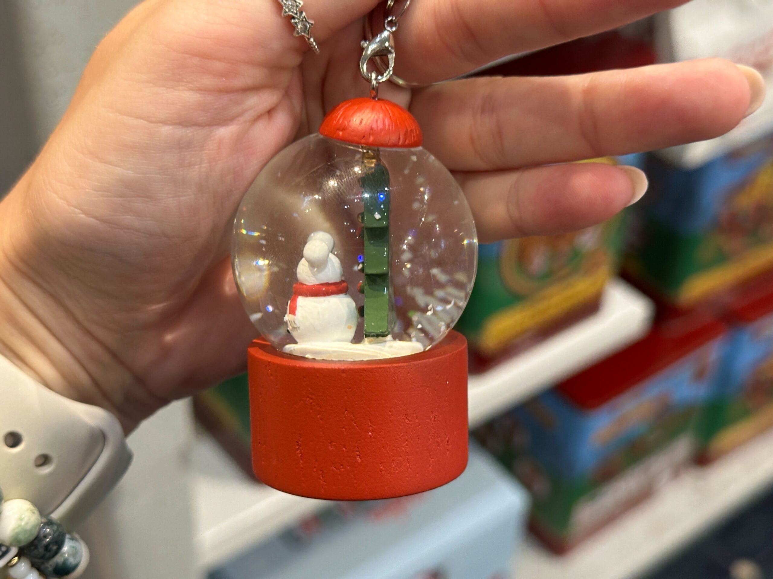 A hand holds a small snow globe keychain with a snowman and a North Pole sign inside on a red base.