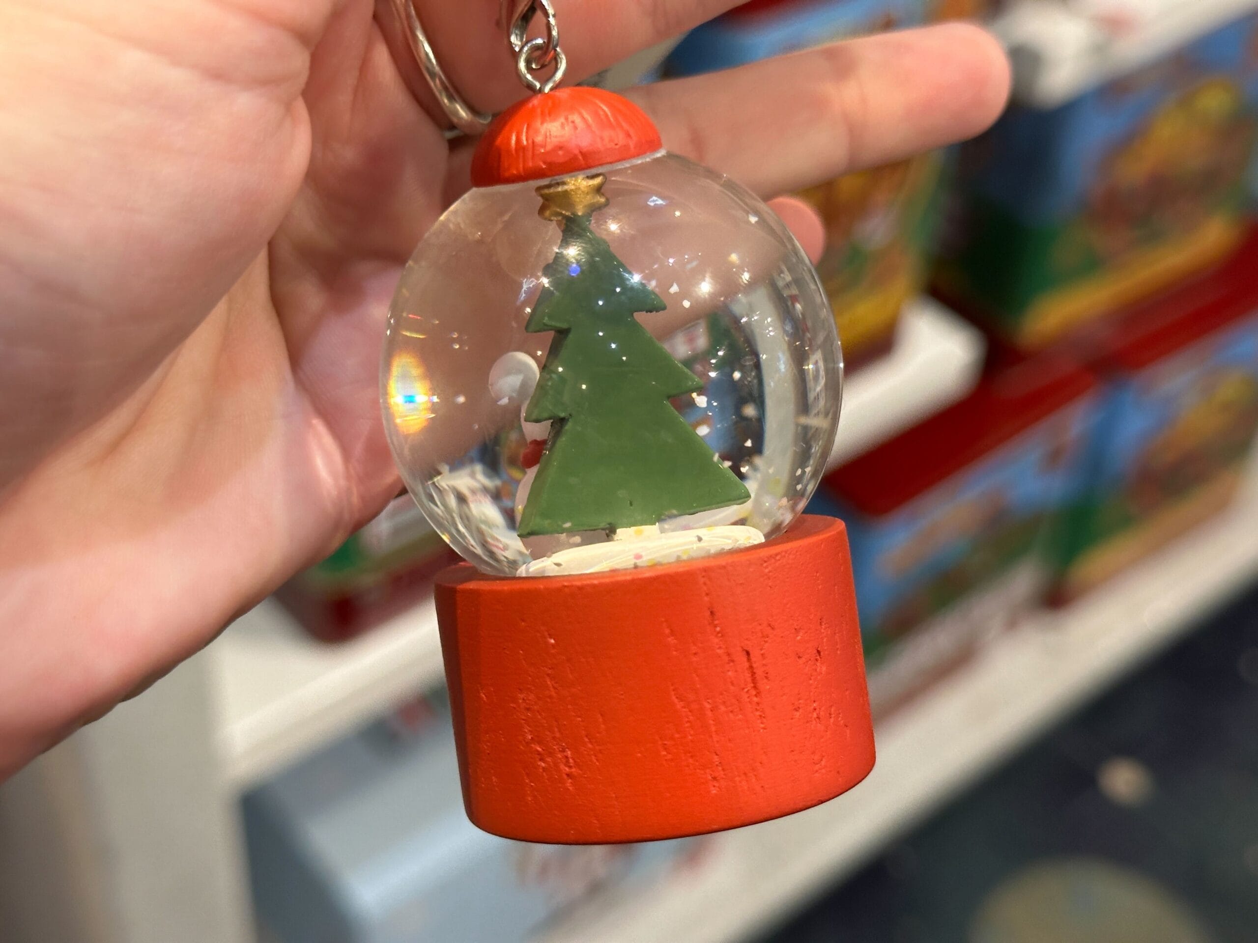 A hand holds a small snow globe ornament with a green Christmas tree inside and a red base.