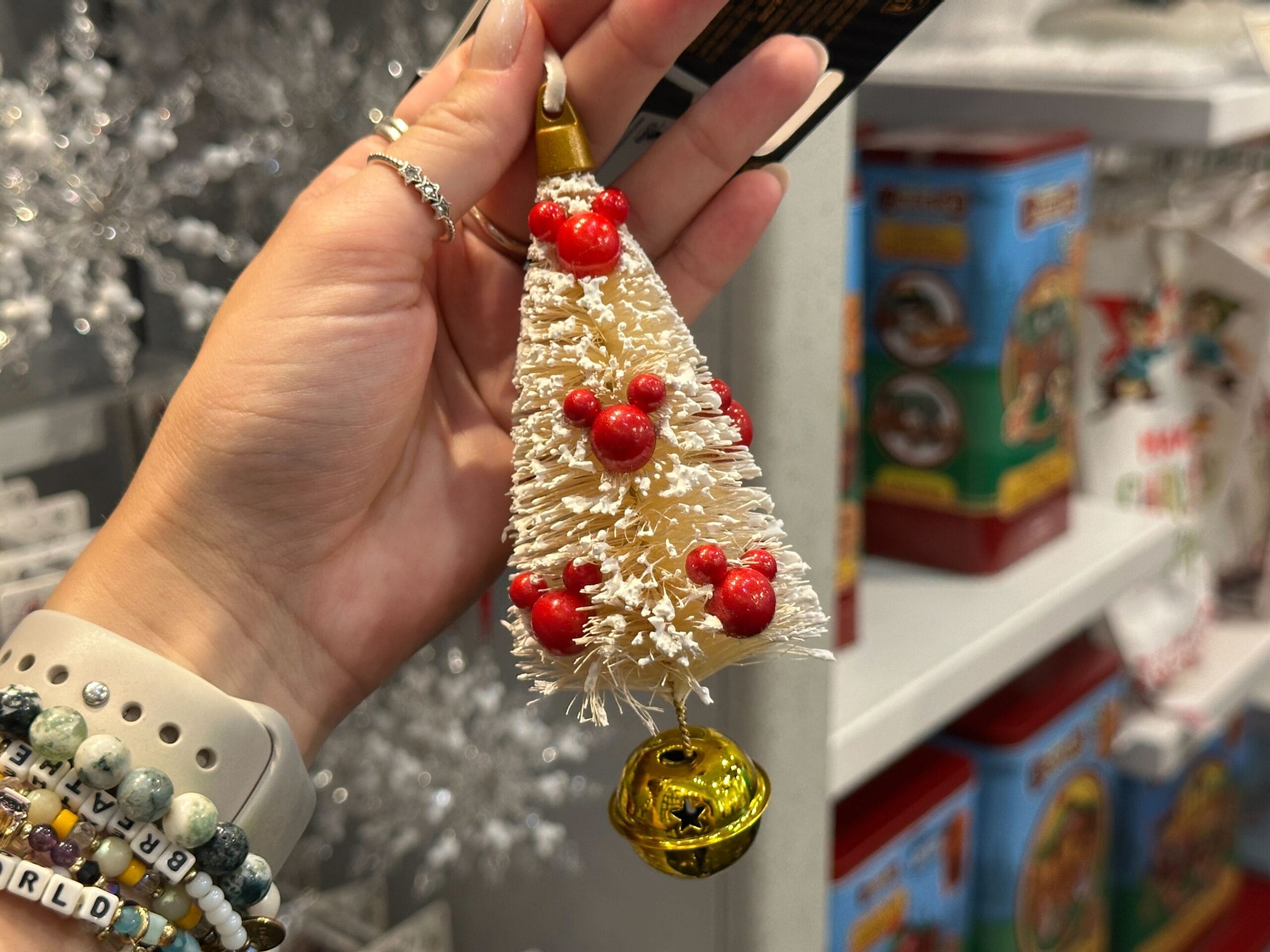 A hand holds a small white Christmas tree ornament with red decorations and a gold bell at the bottom.