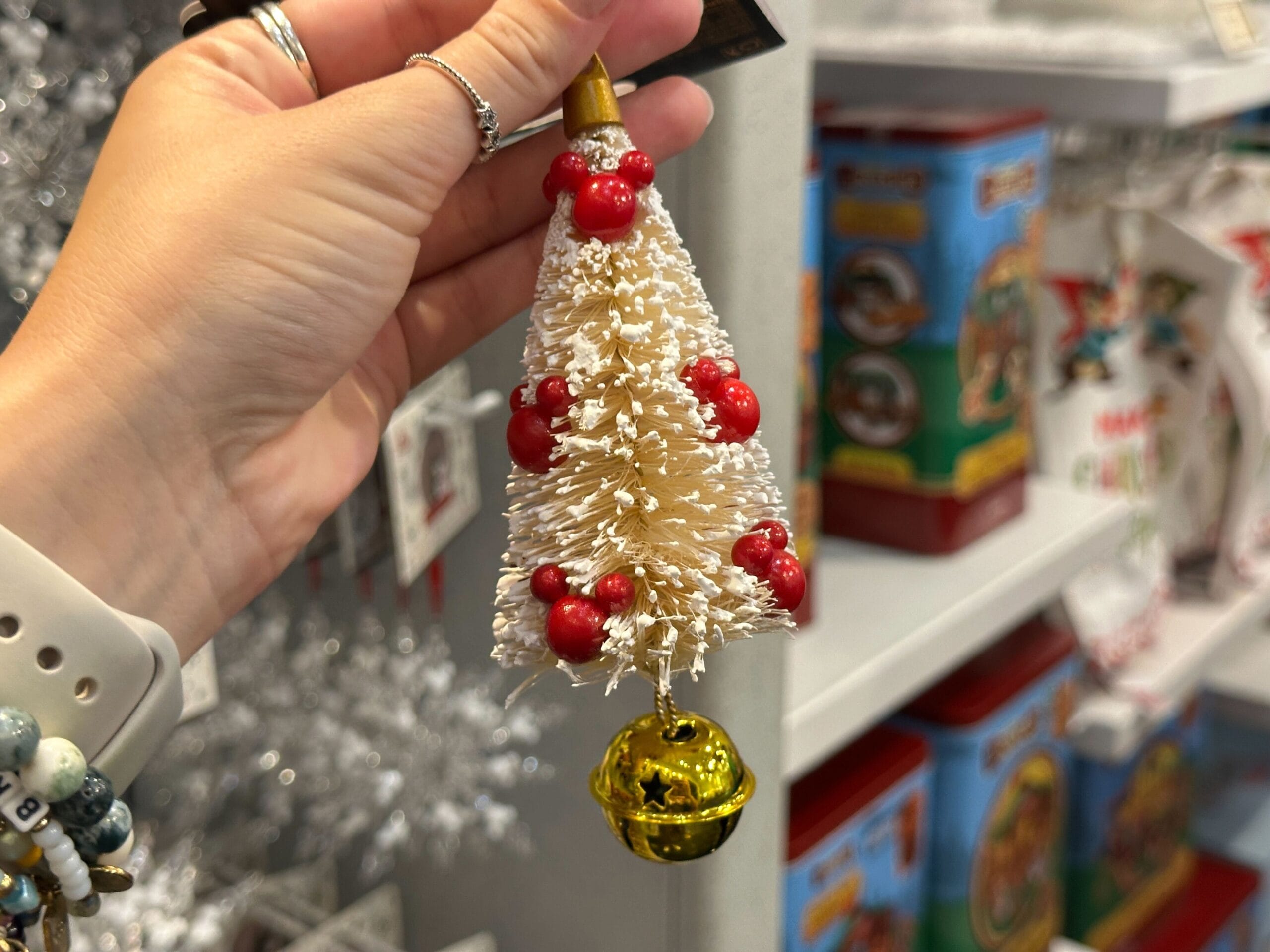 Hand holding a small white Christmas tree ornament with red berries and a gold bell, store shelves in the background.
