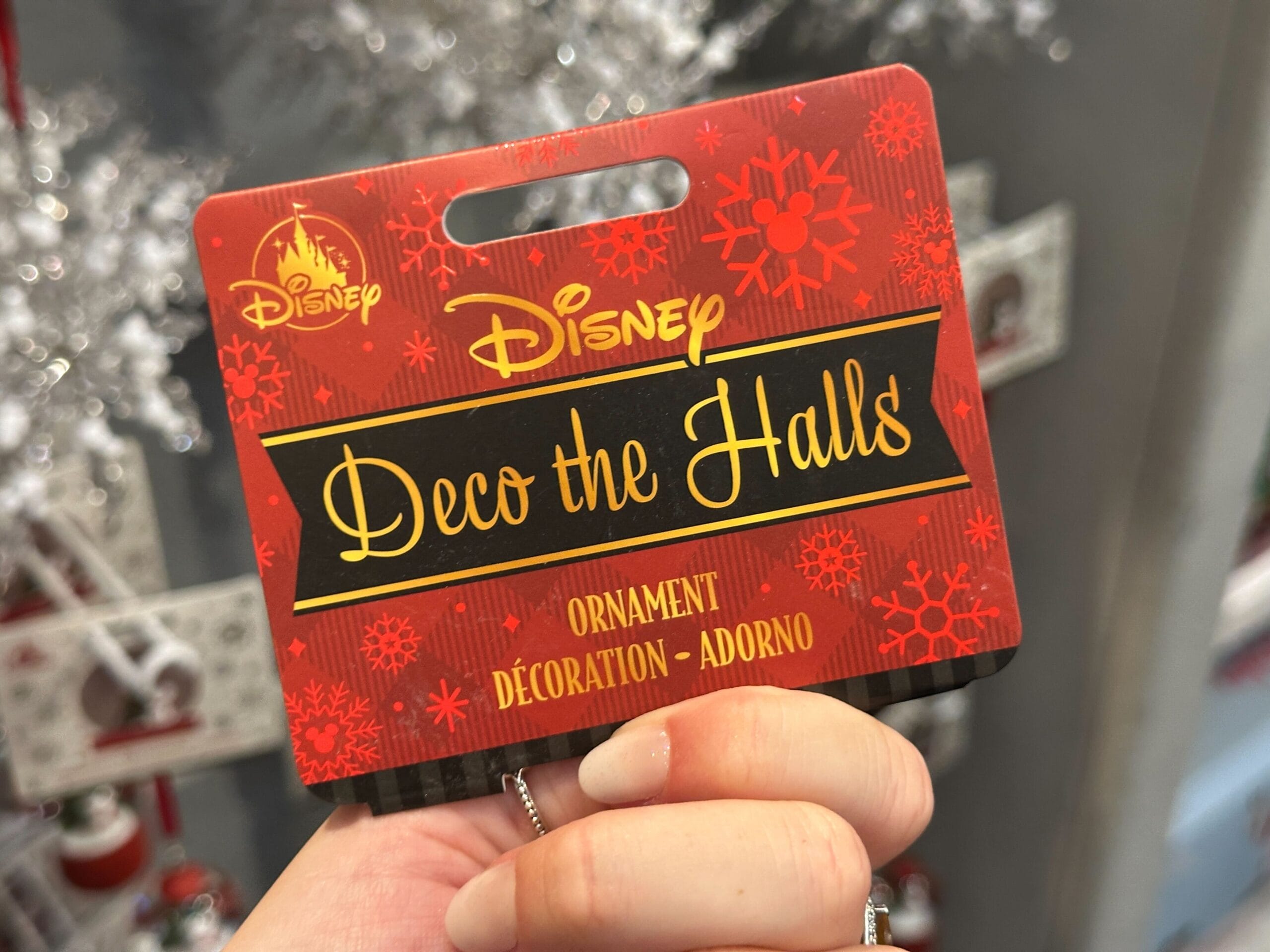 A hand holds a Disney "Deco the Halls" ornament tag decorated with red snowflakes.