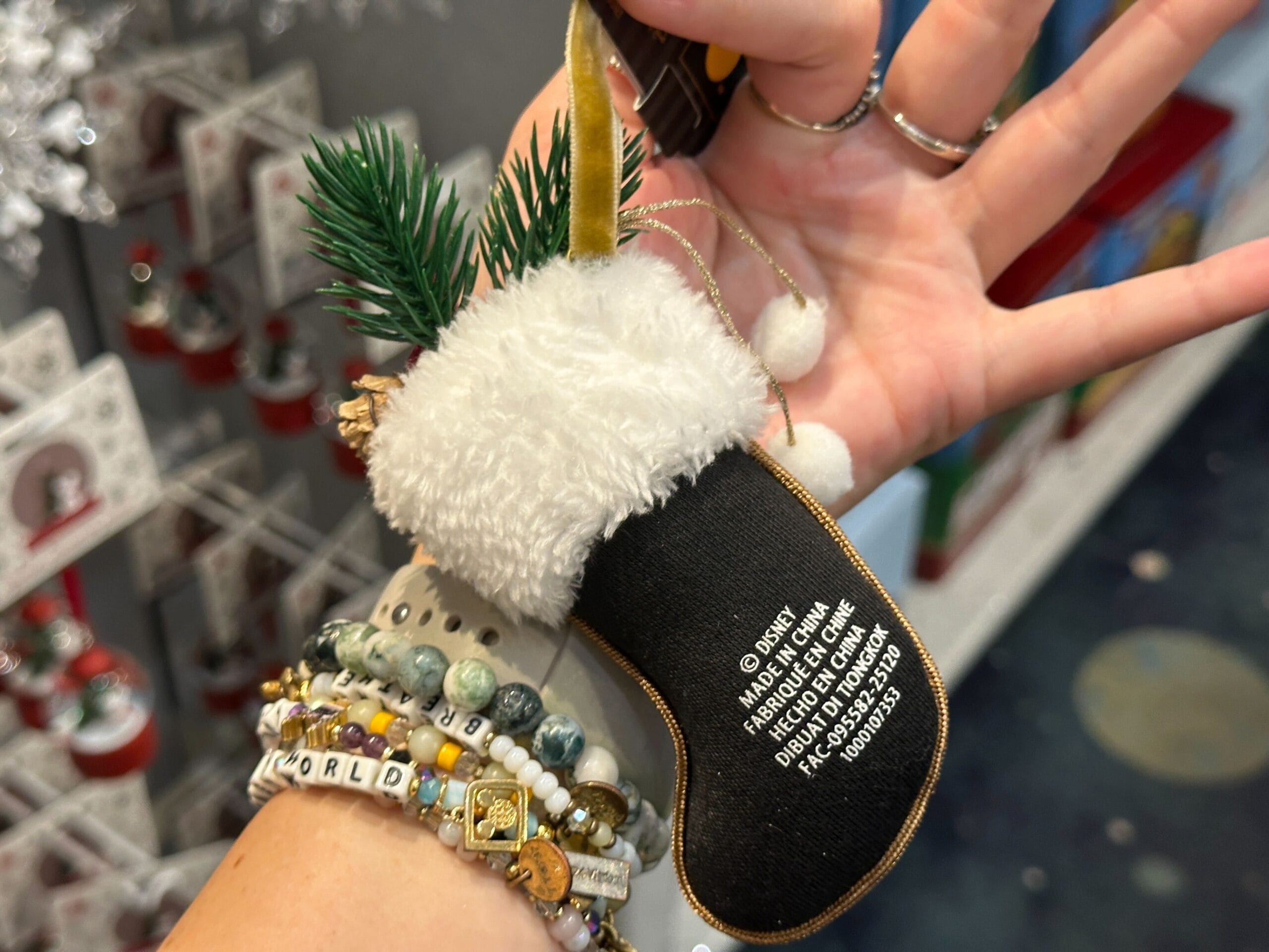 A hand holds a small black Christmas stocking ornament with white fur trim, text, and decorative pine sprigs.