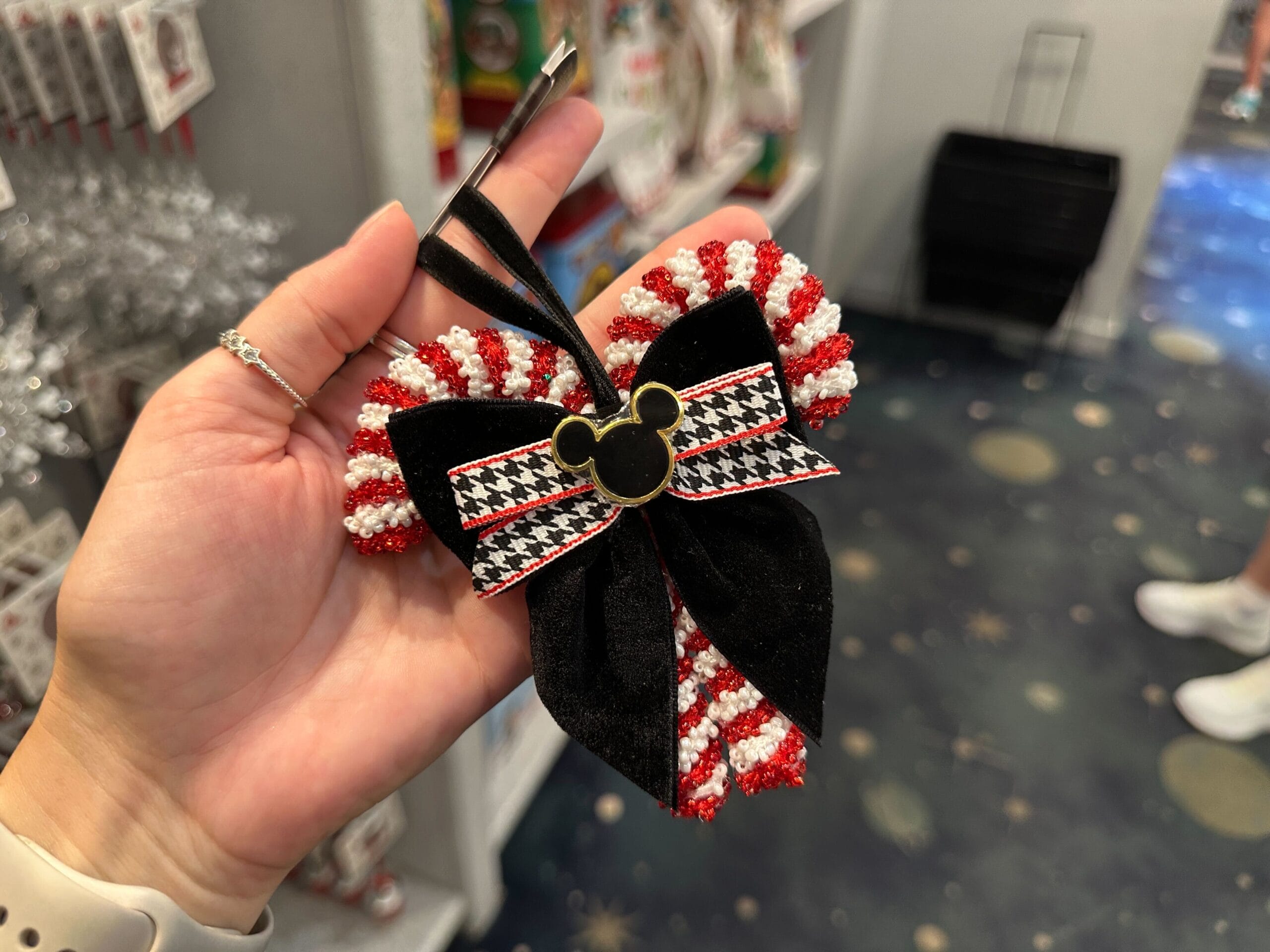 Hand holding a festive bow ornament with red and white stripes, black velvet, and a Mickey Mouse emblem in the center.