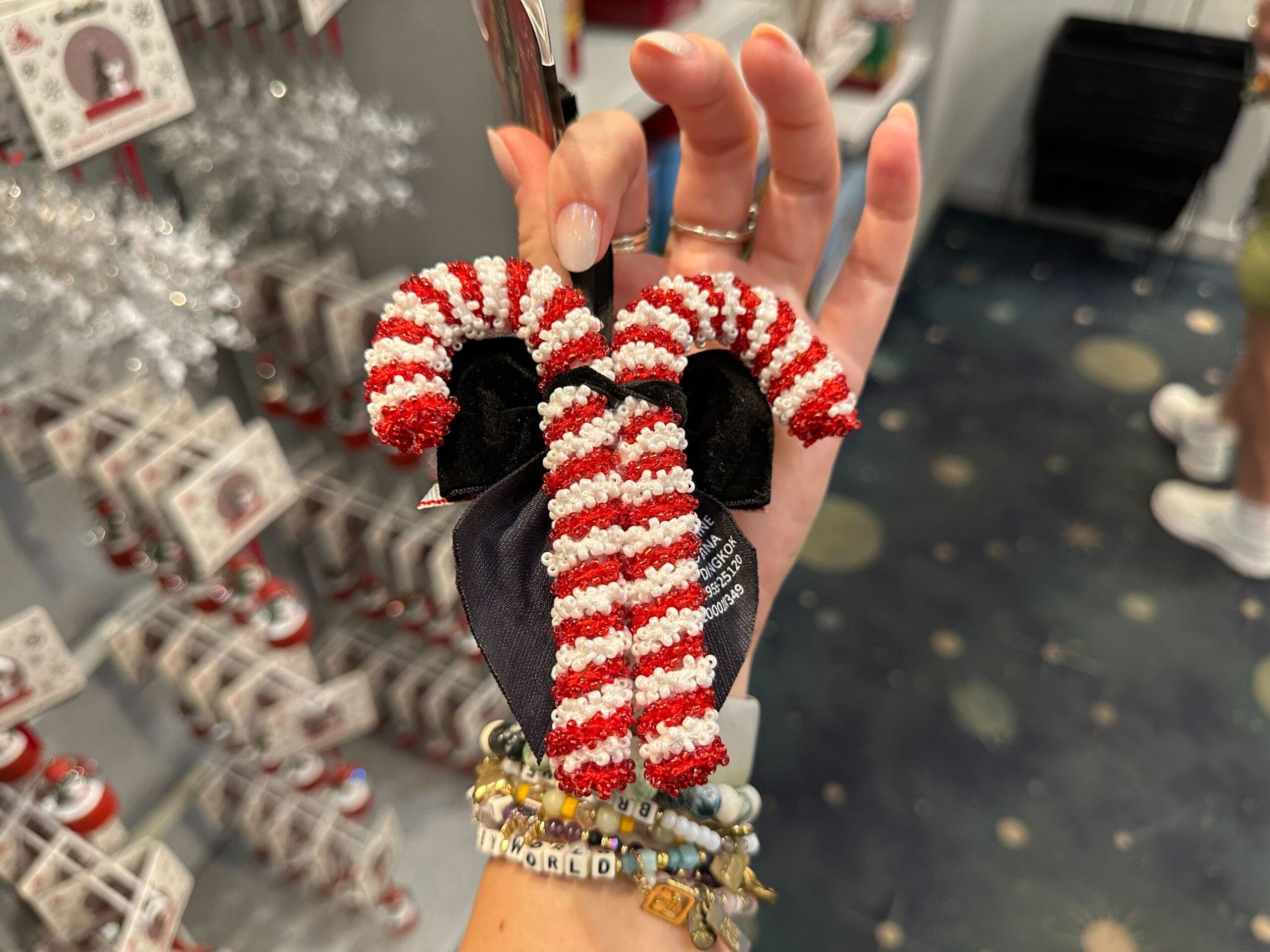 A hand holds a beaded candy cane ornament with a black bow, surrounded by holiday decorations in a store.