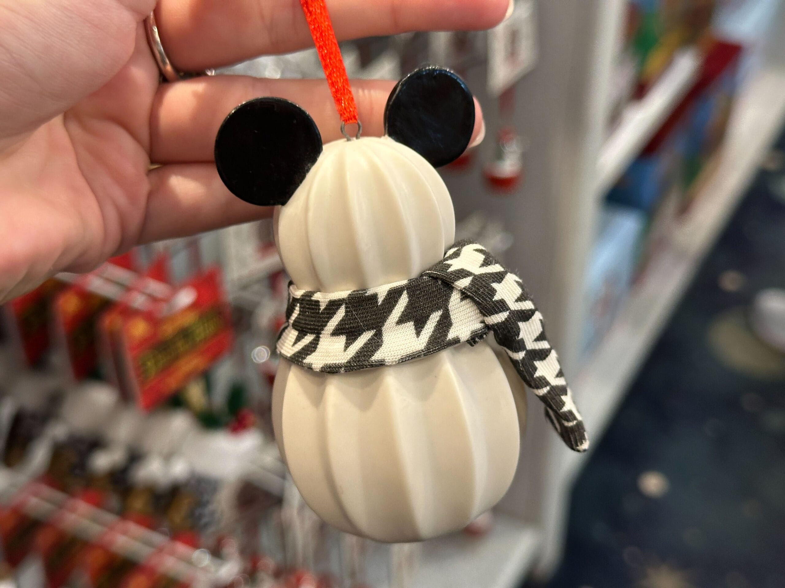A hand holds a snowman ornament with Mickey Mouse ears and a black-and-white houndstooth scarf.