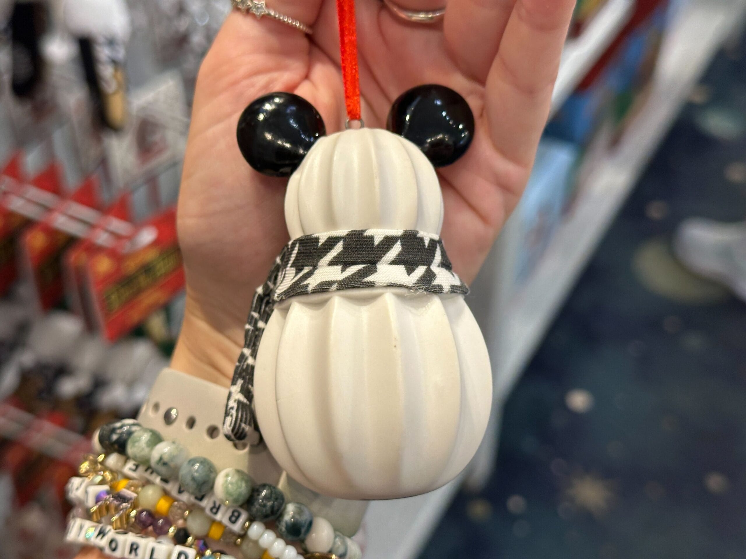 A hand holds a white snowman ornament with black ears and a houndstooth scarf in a store.