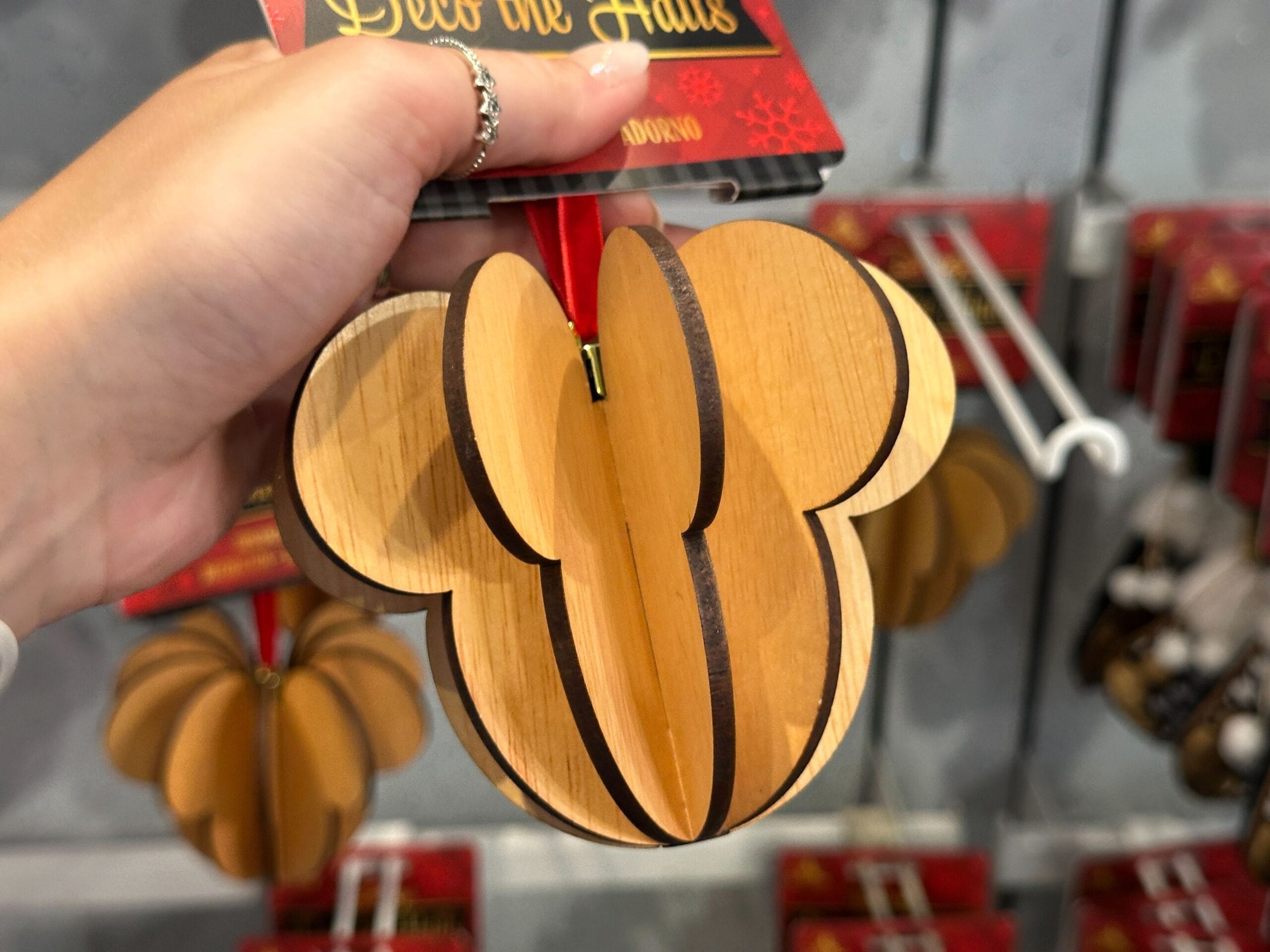 A hand holds a wooden ornament shaped like Mickey Mouse’s head with a red ribbon for hanging.