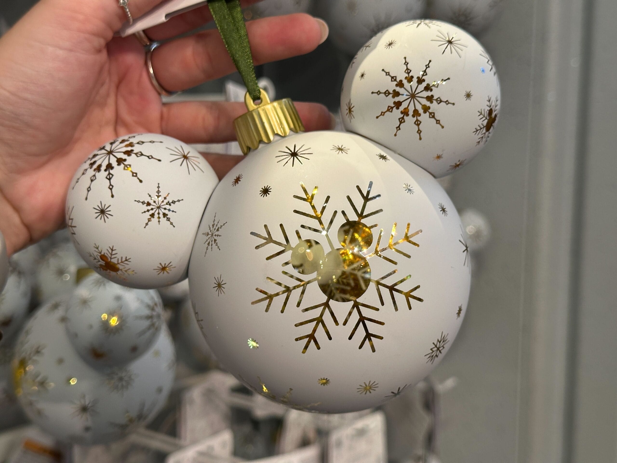 Hand holding a white Mickey Mouse-shaped ornament decorated with gold snowflakes and a green ribbon.