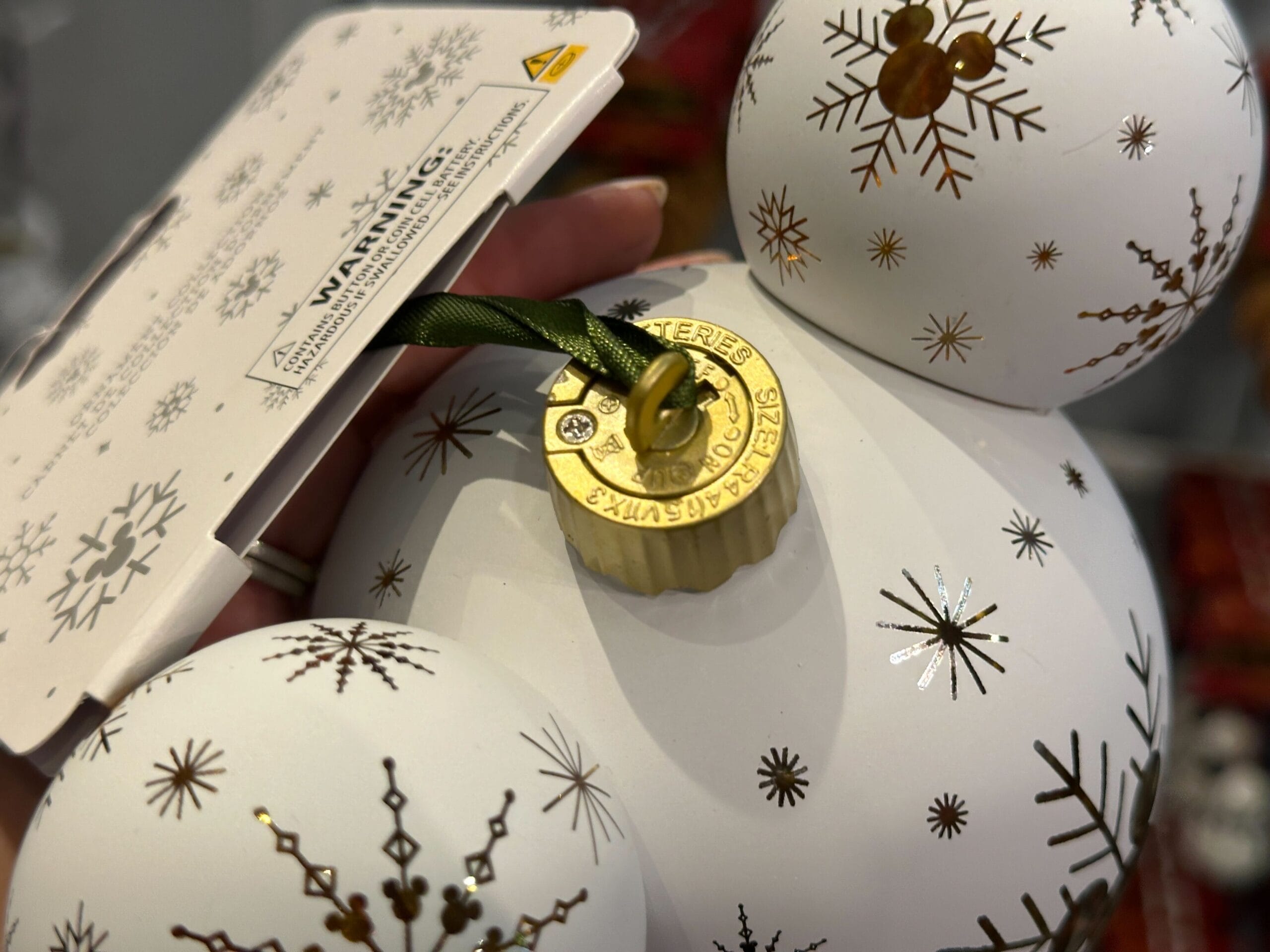 Close-up of white Christmas ornaments with gold snowflake patterns and a gold cap, held by a hand with a warning tag.