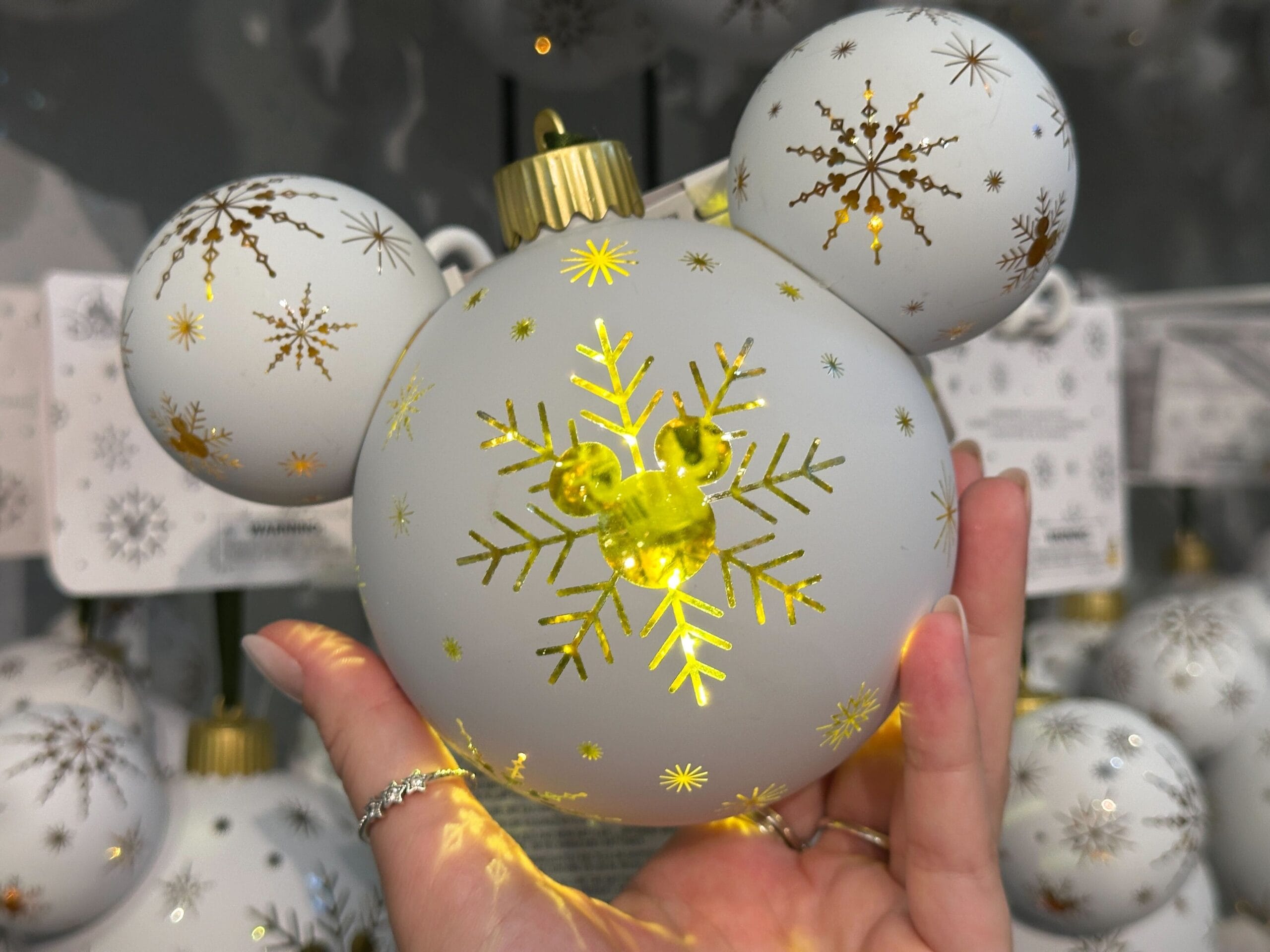 A hand holds a Disney-themed white ornament with gold snowflakes and Mickey Mouse ears, glowing with yellow light.