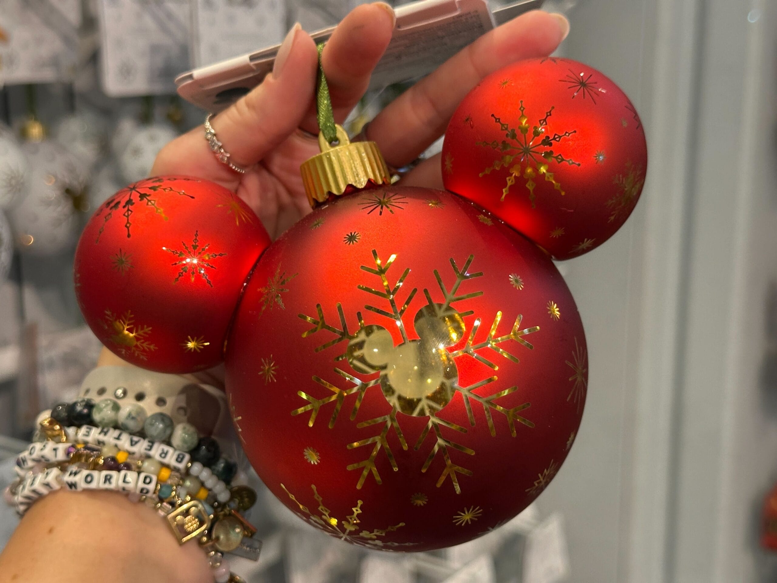 A hand holds a red Mickey Mouse-shaped ornament decorated with gold snowflakes and a gold Mickey silhouette.