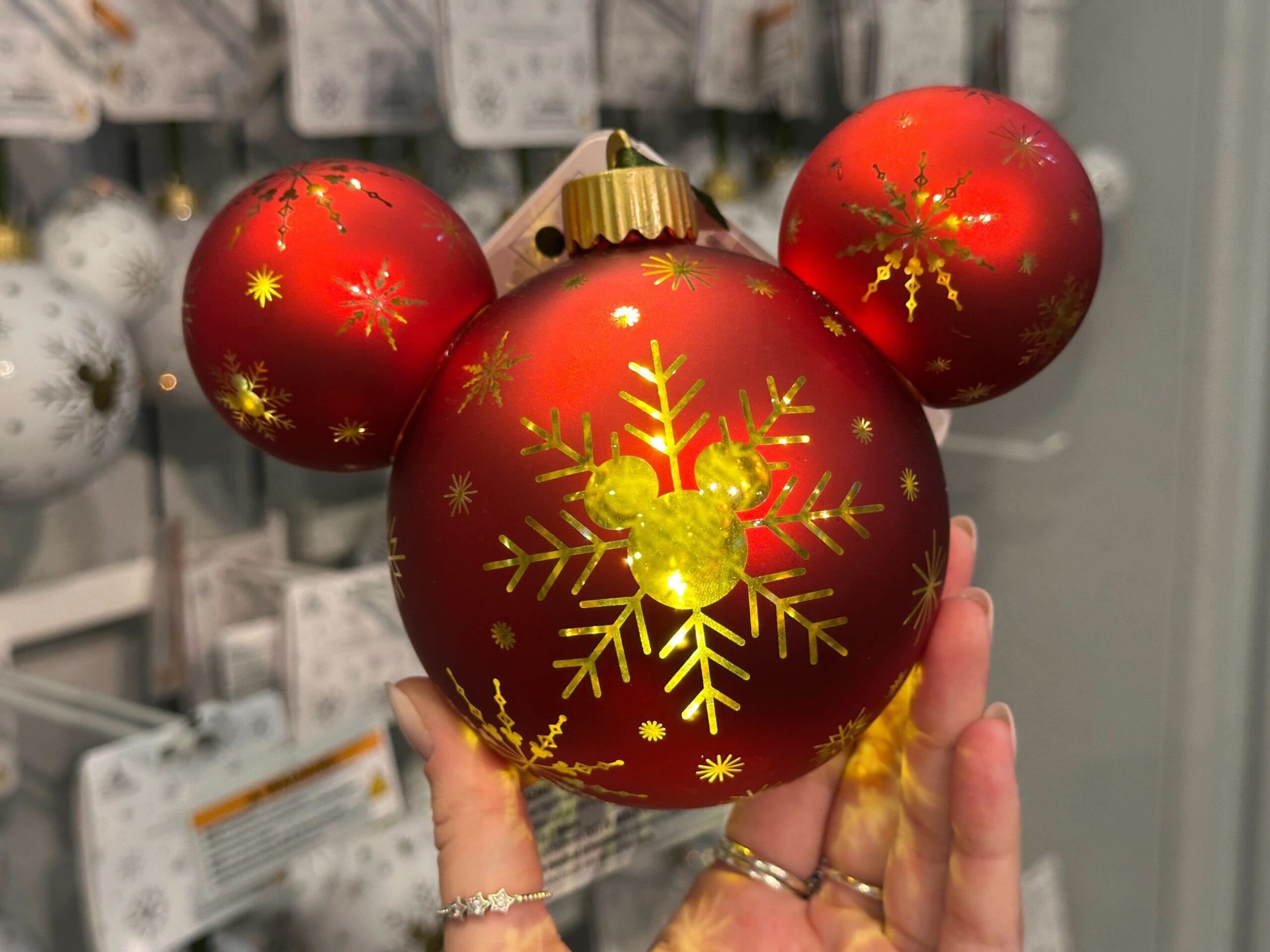 A hand holds a red Mickey Mouse-shaped Christmas ornament with gold snowflakes and ears.