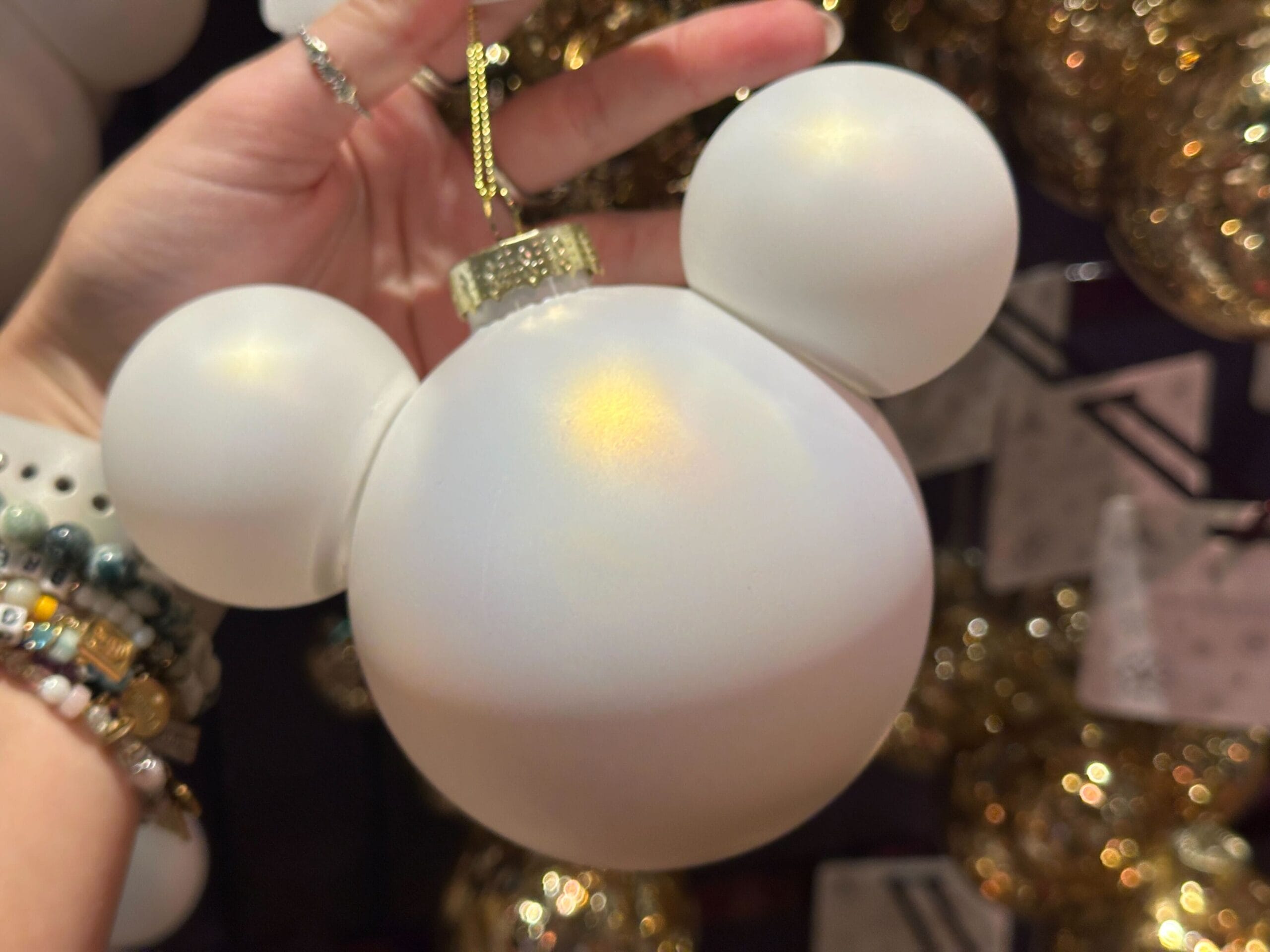 A hand holds a white Mickey Mouse-shaped ornament with a gold cap, surrounded by glittery gold ornaments.