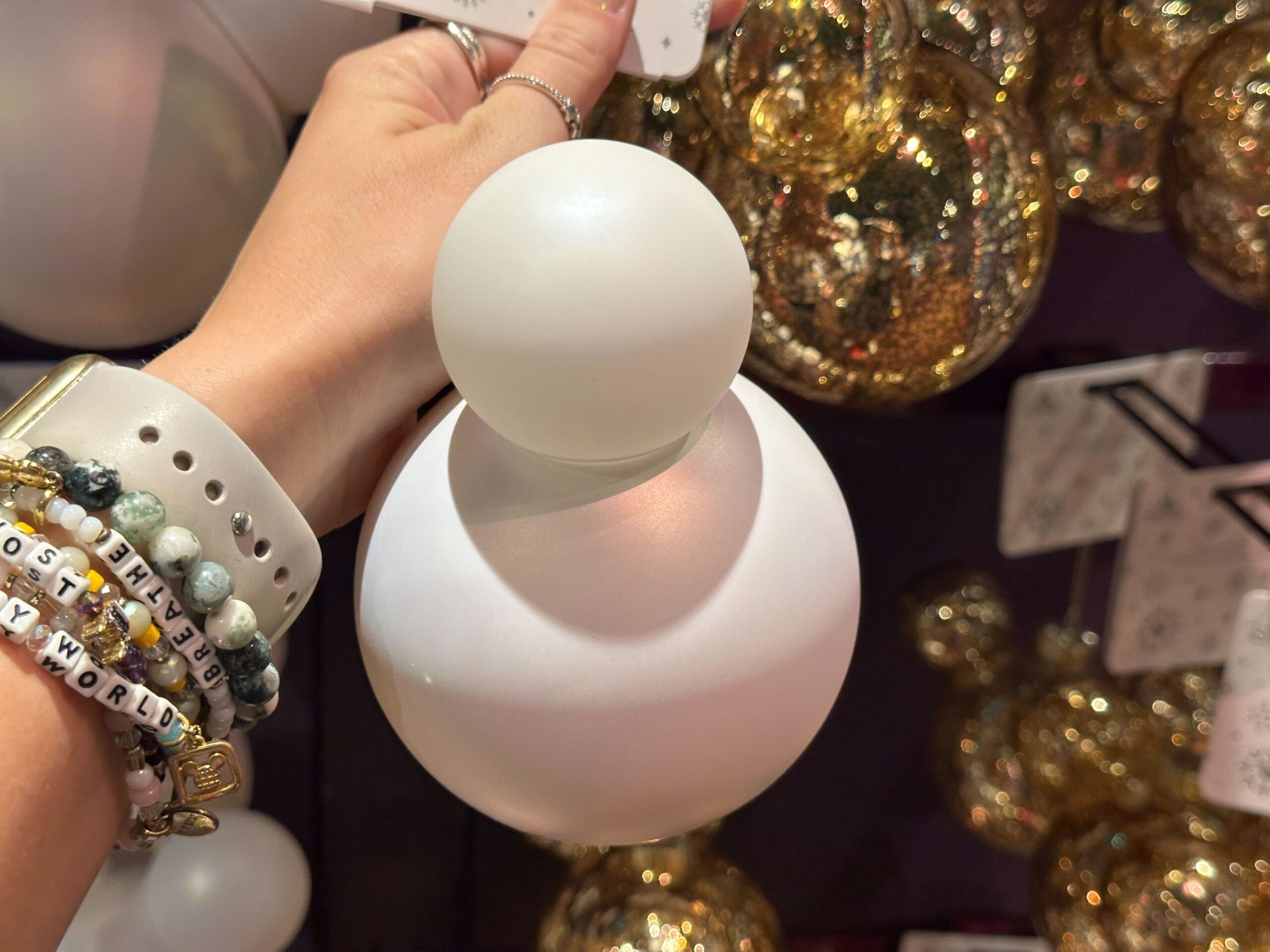 A hand holding a white ornament with two spheres, surrounded by shiny gold ornaments in the background.