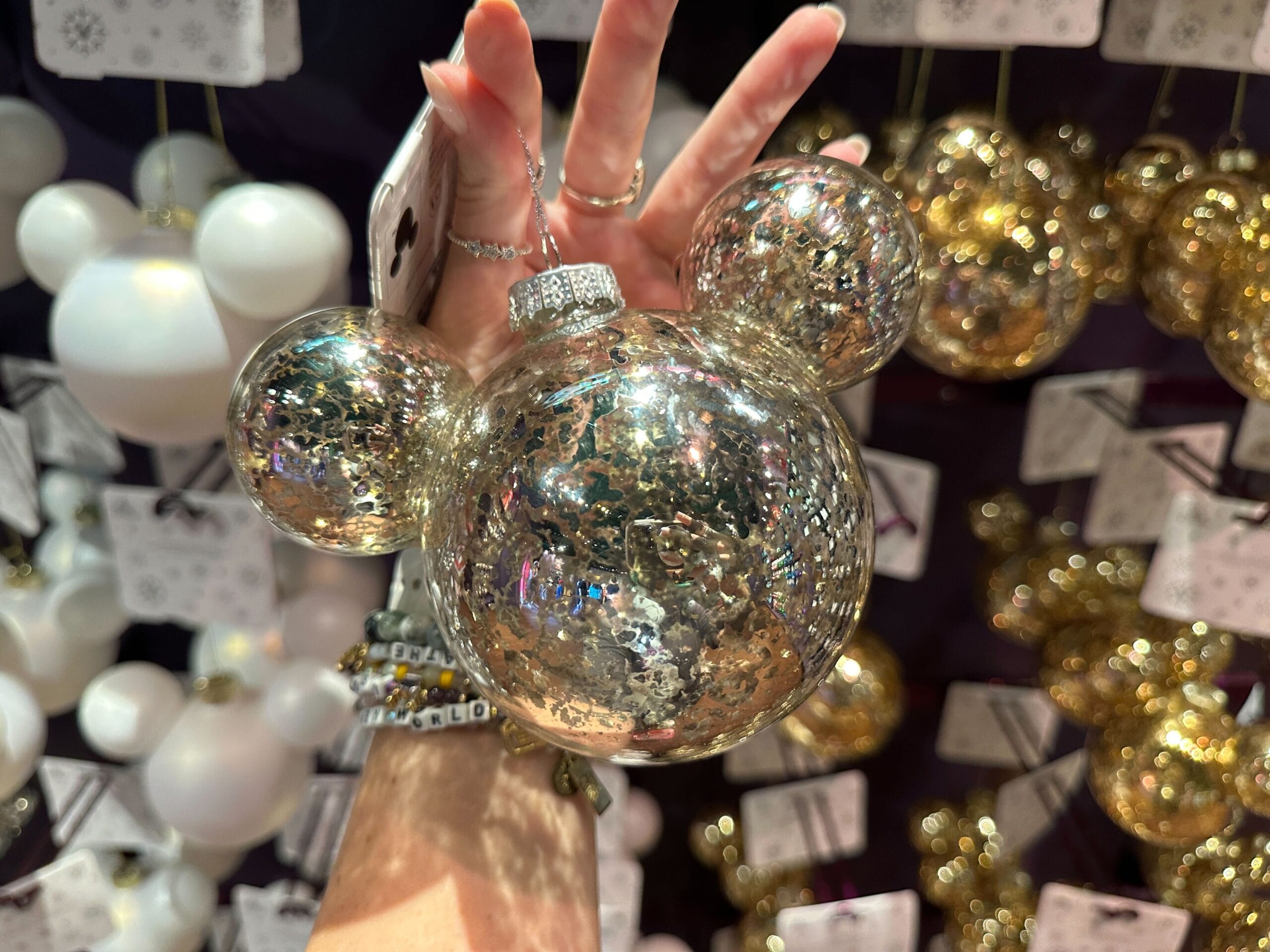 A hand holds a sparkly Mickey Mouse-shaped ornament among other shiny ornaments on display.