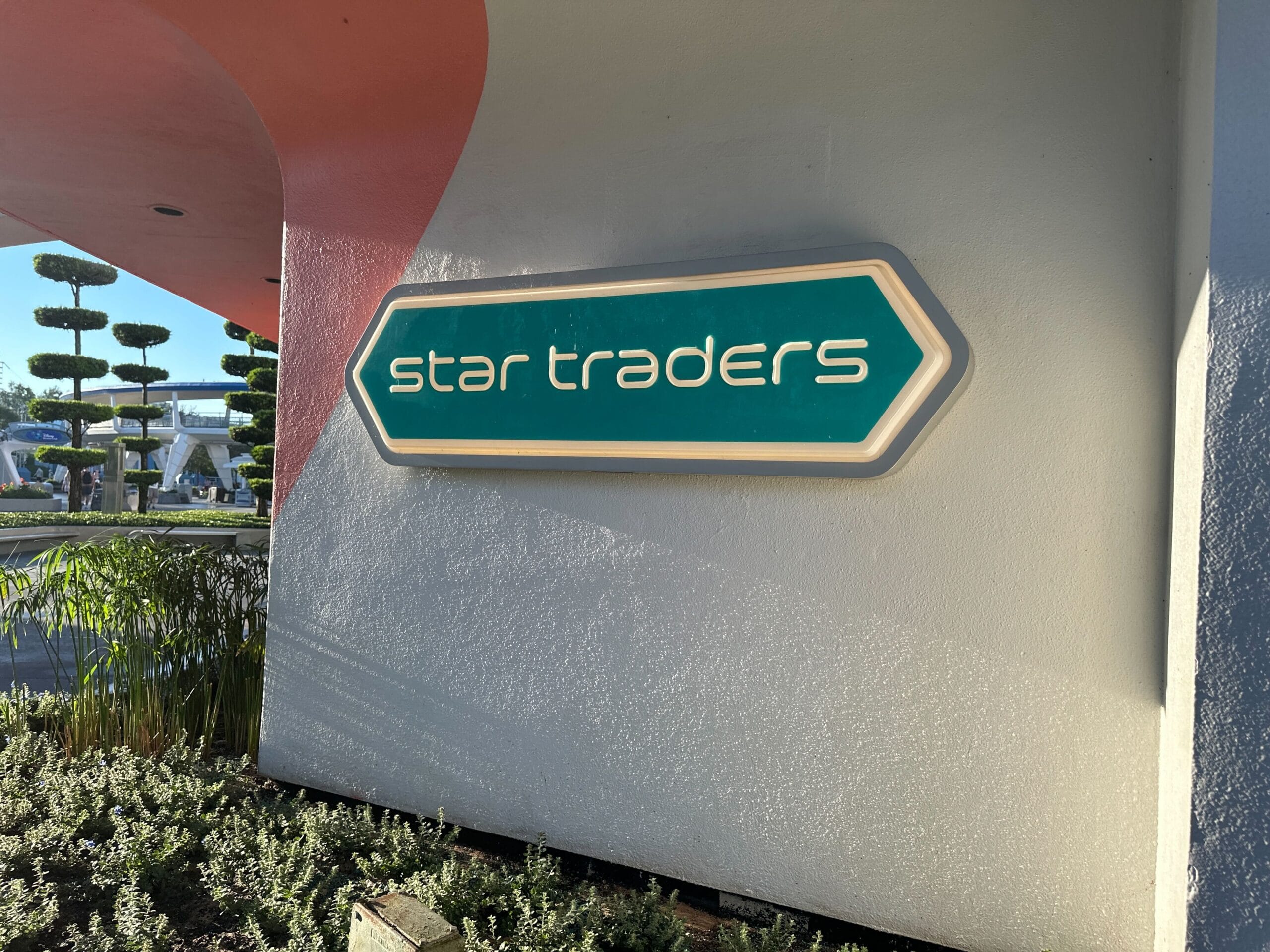 A teal Star Traders sign is mounted on a white wall next to shrubs and geometric trees.