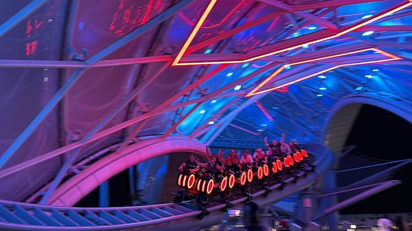 People ride a futuristic, neon-lit roller coaster with a TRON Nighttime Overlay under a glowing blue and red canopy.