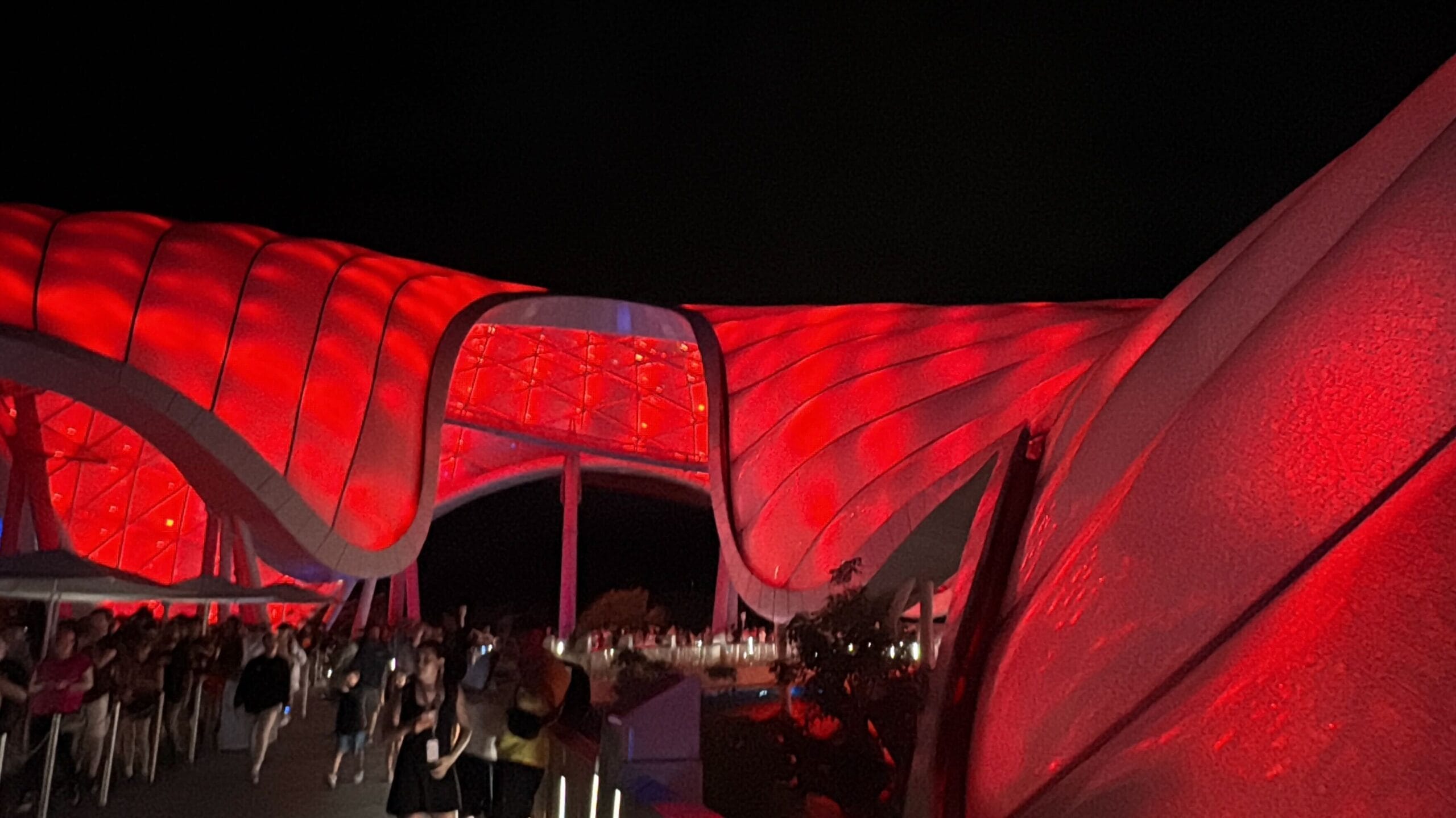 A crowd walks under a large, futuristic structure with a TRON Nighttime Overlay illuminated by red lights at night.