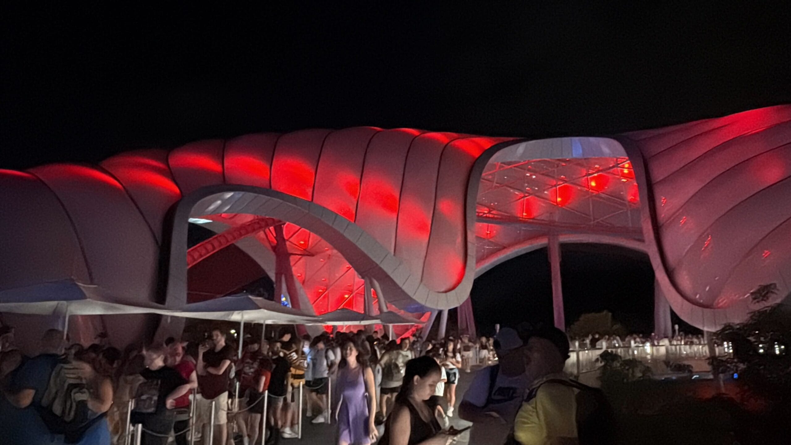 People stand in line at night under a wavy, futuristic structure with a TRON Nighttime Overlay and glowing red lights.