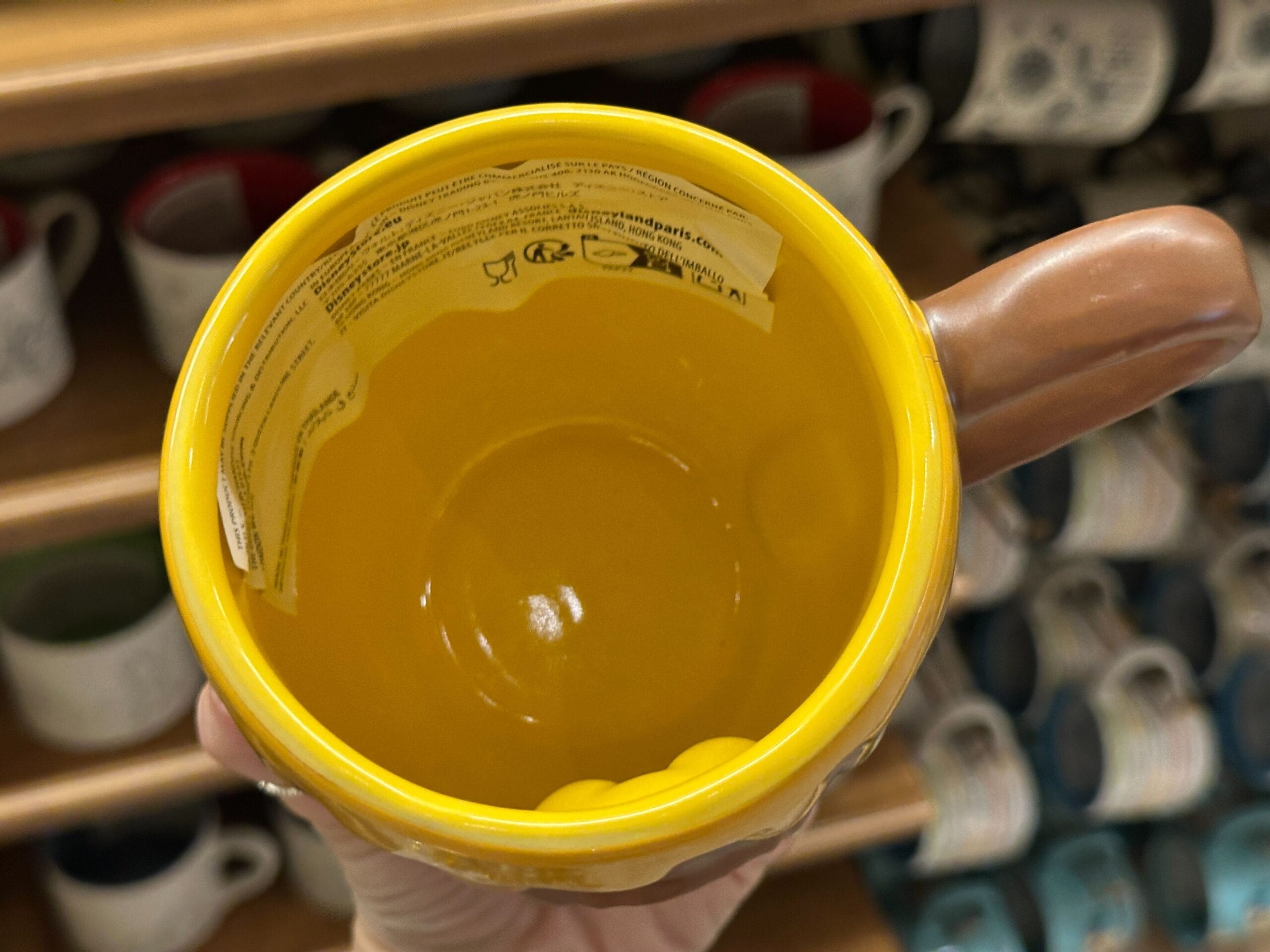 A hand holds a yellow ceramic mug with a label inside, in front of shelves filled with assorted mugs.