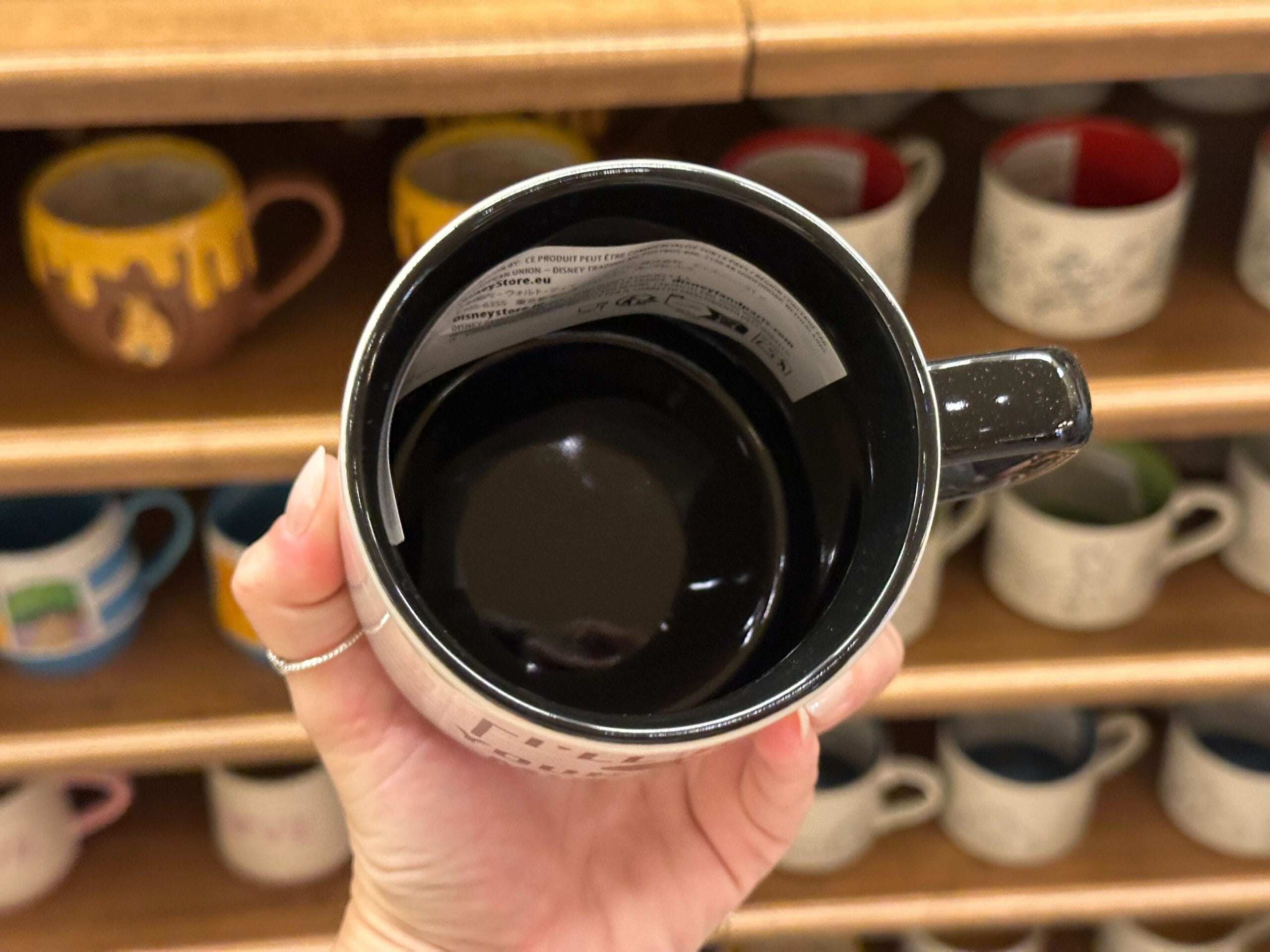 A hand holds a black coffee mug over shelves filled with various colorful mugs in a store.