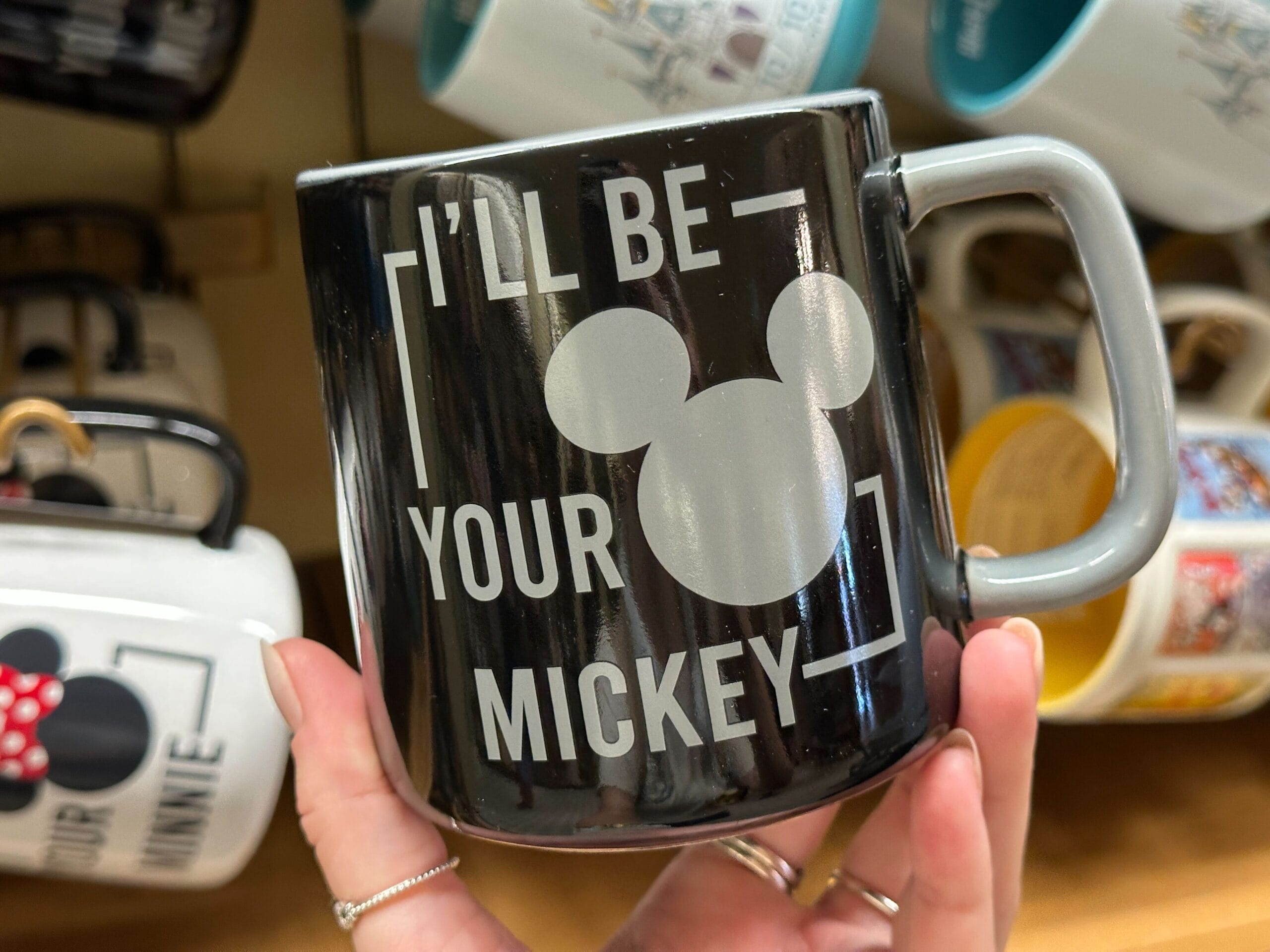 A hand holds a black mug with a Mickey Mouse silhouette and the text "I'LL BE YOUR MICKEY.
