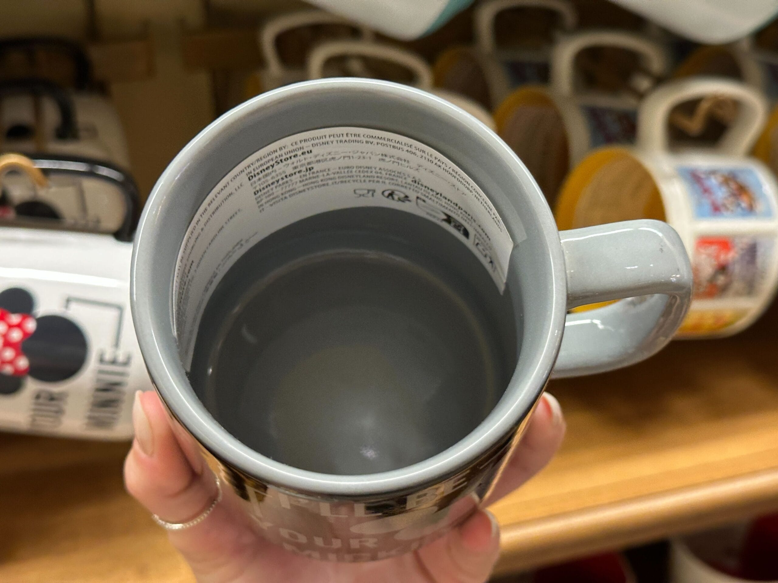 A hand holds a gray mug filled with water; other decorative mugs are displayed on a shelf in the background.