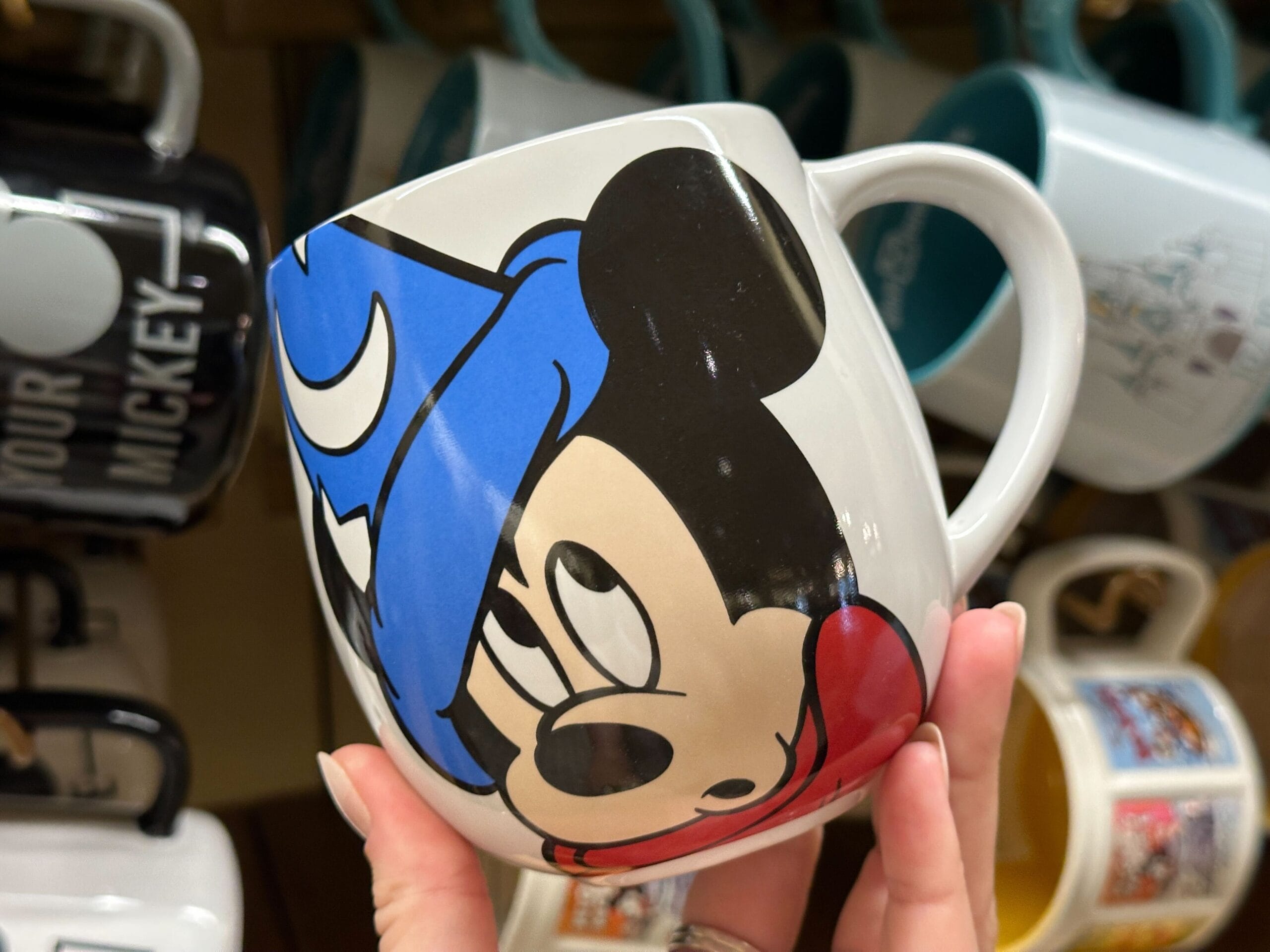 A hand holds a mug featuring Mickey Mouse in a blue wizard hat, with other mugs displayed in the background.