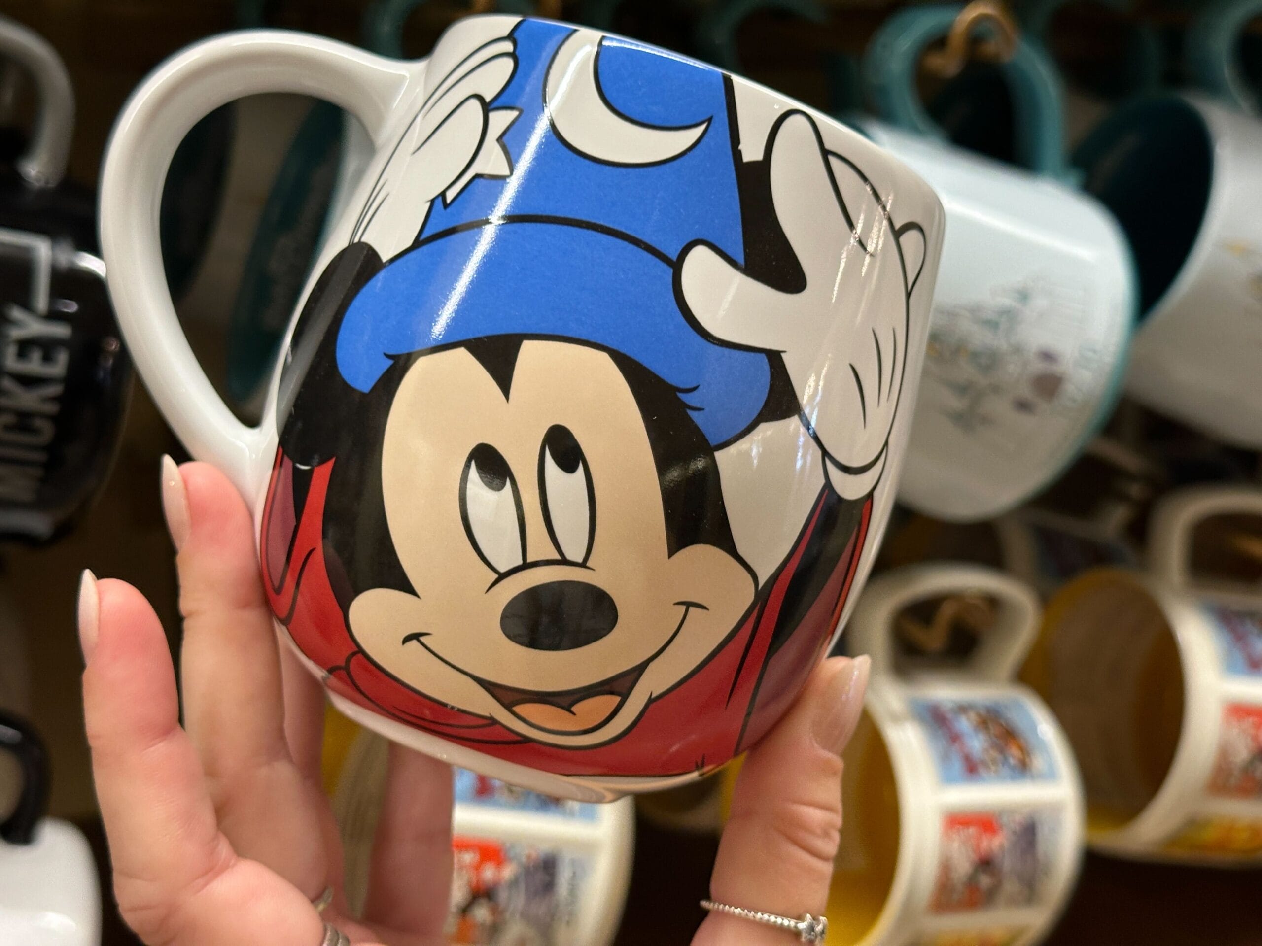 Hand holding a mug featuring Minnie Mouse wearing a blue sorcerer's hat; mugs are displayed in the background.