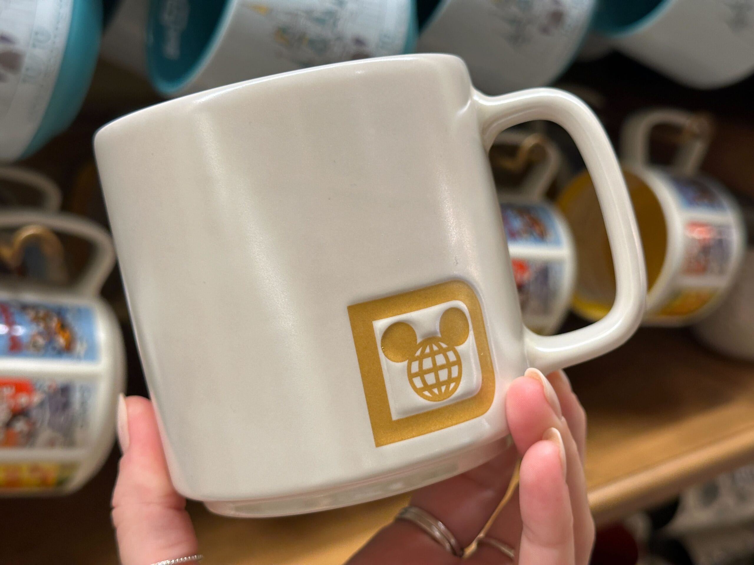 A white mug with a gold square logo featuring a Mickey Mouse head and a globe, held by a hand in a store.