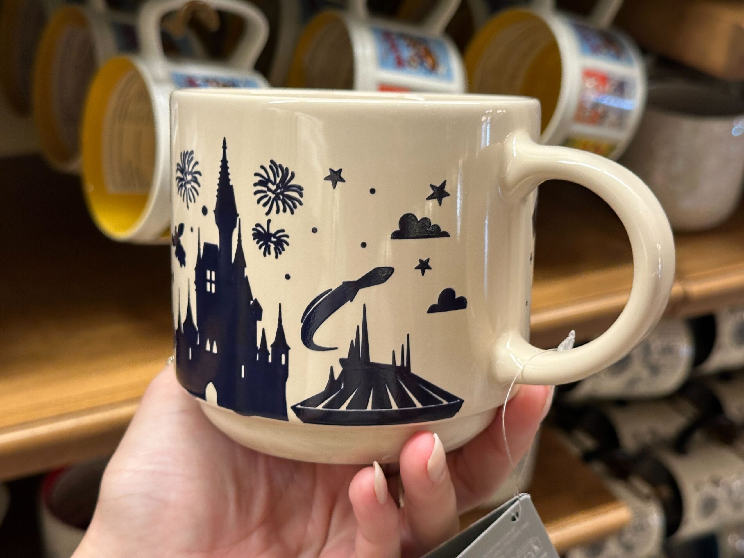 Beige mug with black castle, fireworks, stars, rocket ship, and Mickey silhouettes, held in a person's hand.