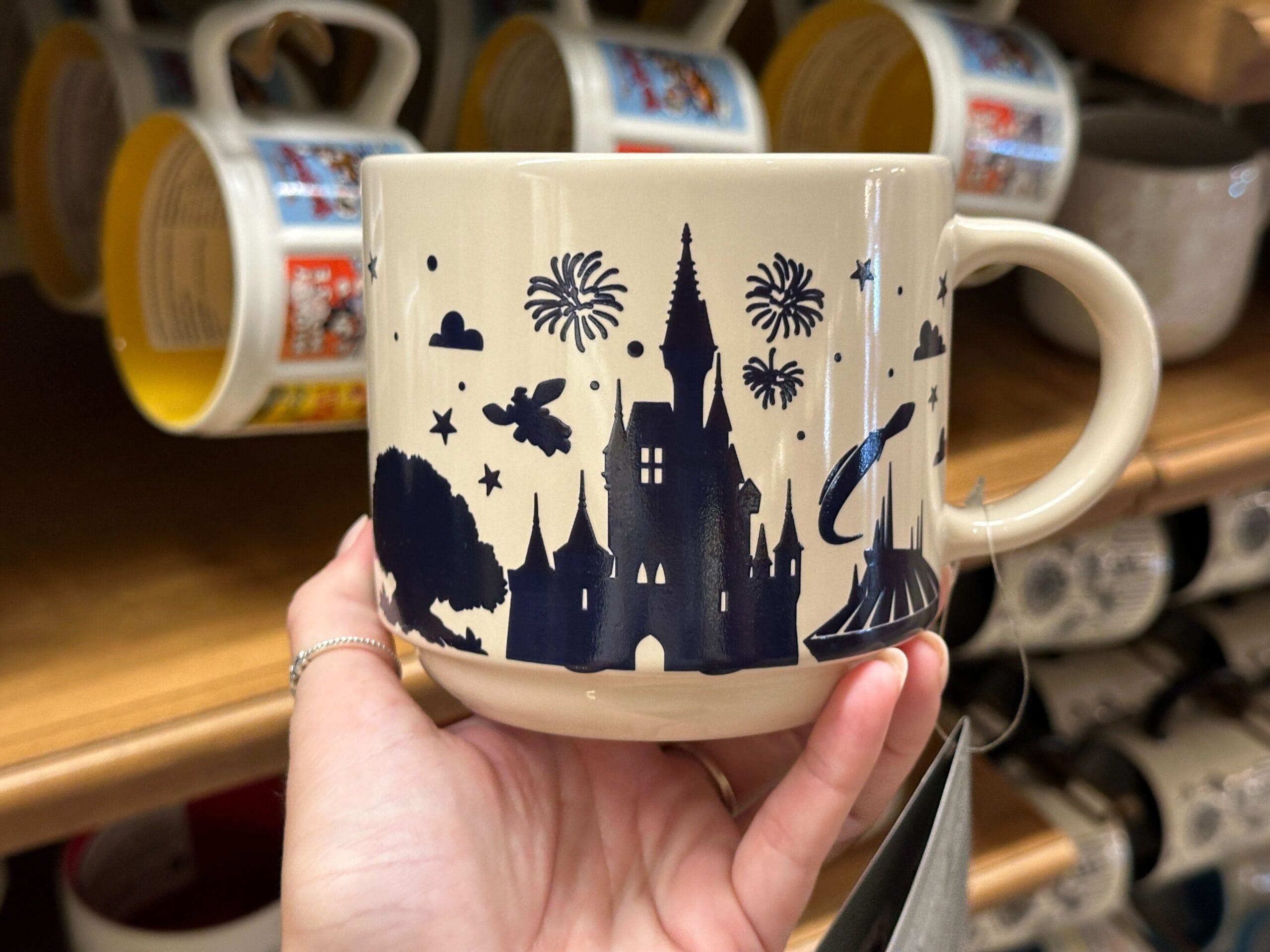 Beige mug with navy blue silhouettes of a castle, fireworks, and Mickey, held in a hand in a store display.
