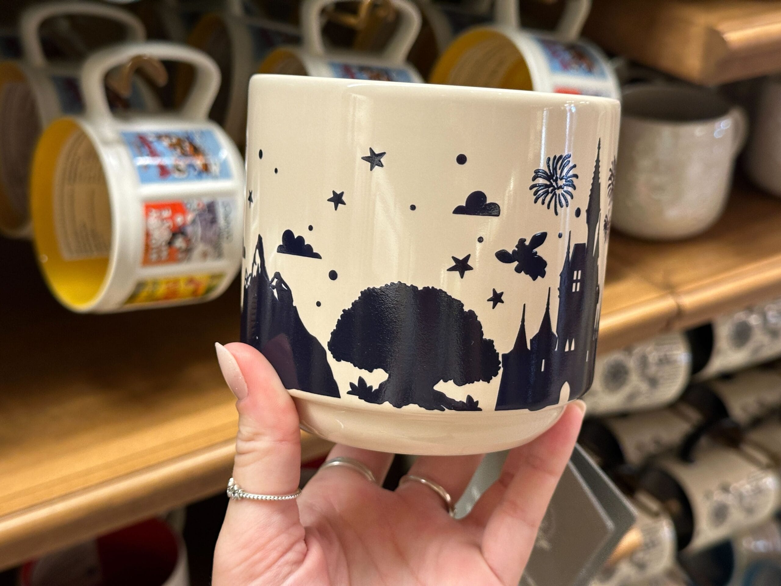 A hand holds a mug featuring Mickey, black tree silhouettes, stars, fireworks, and a castle on a cream background.