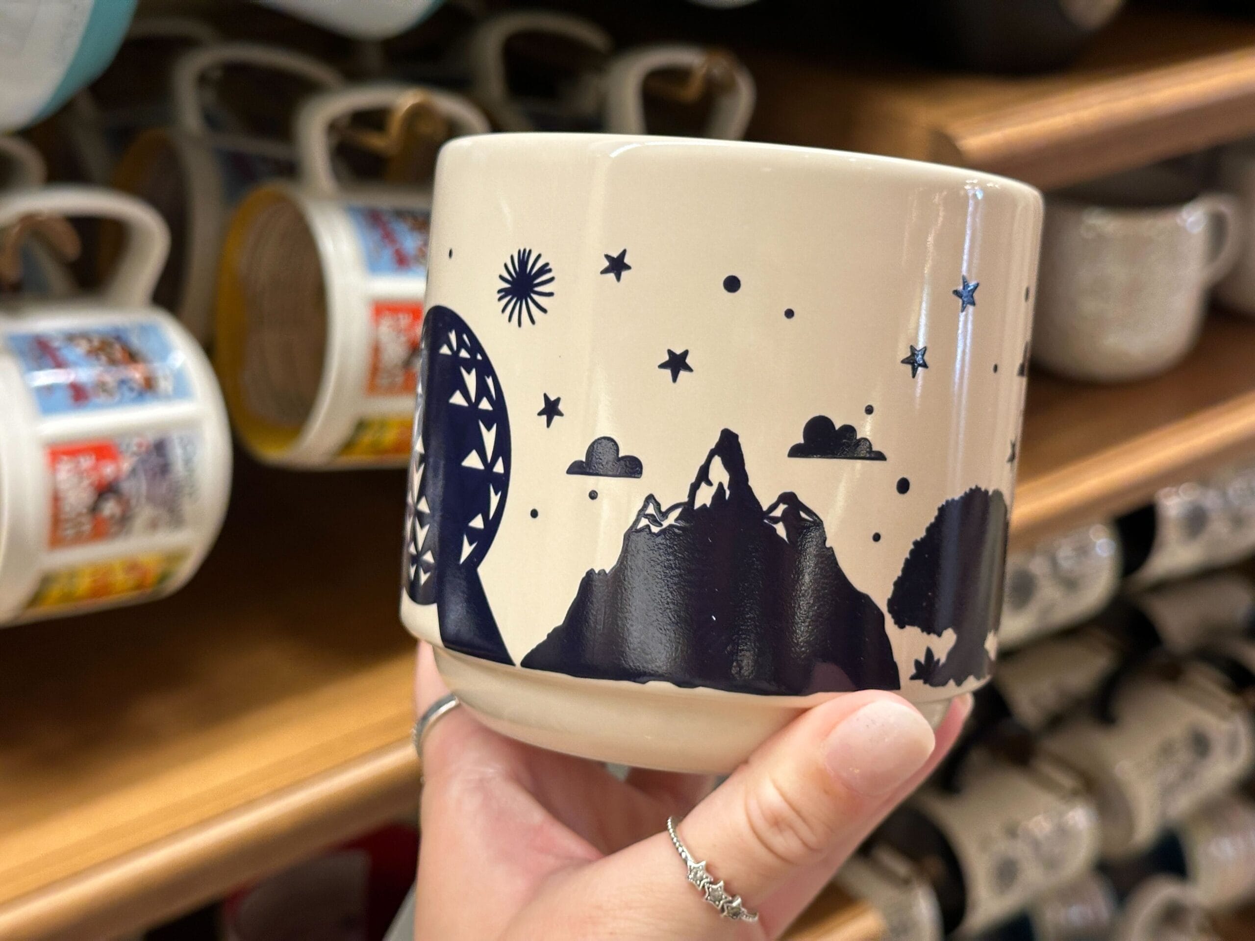 A hand holds a mug featuring Mickey, a mountain, tree, moon, stars, and clouds in a dark blue design.