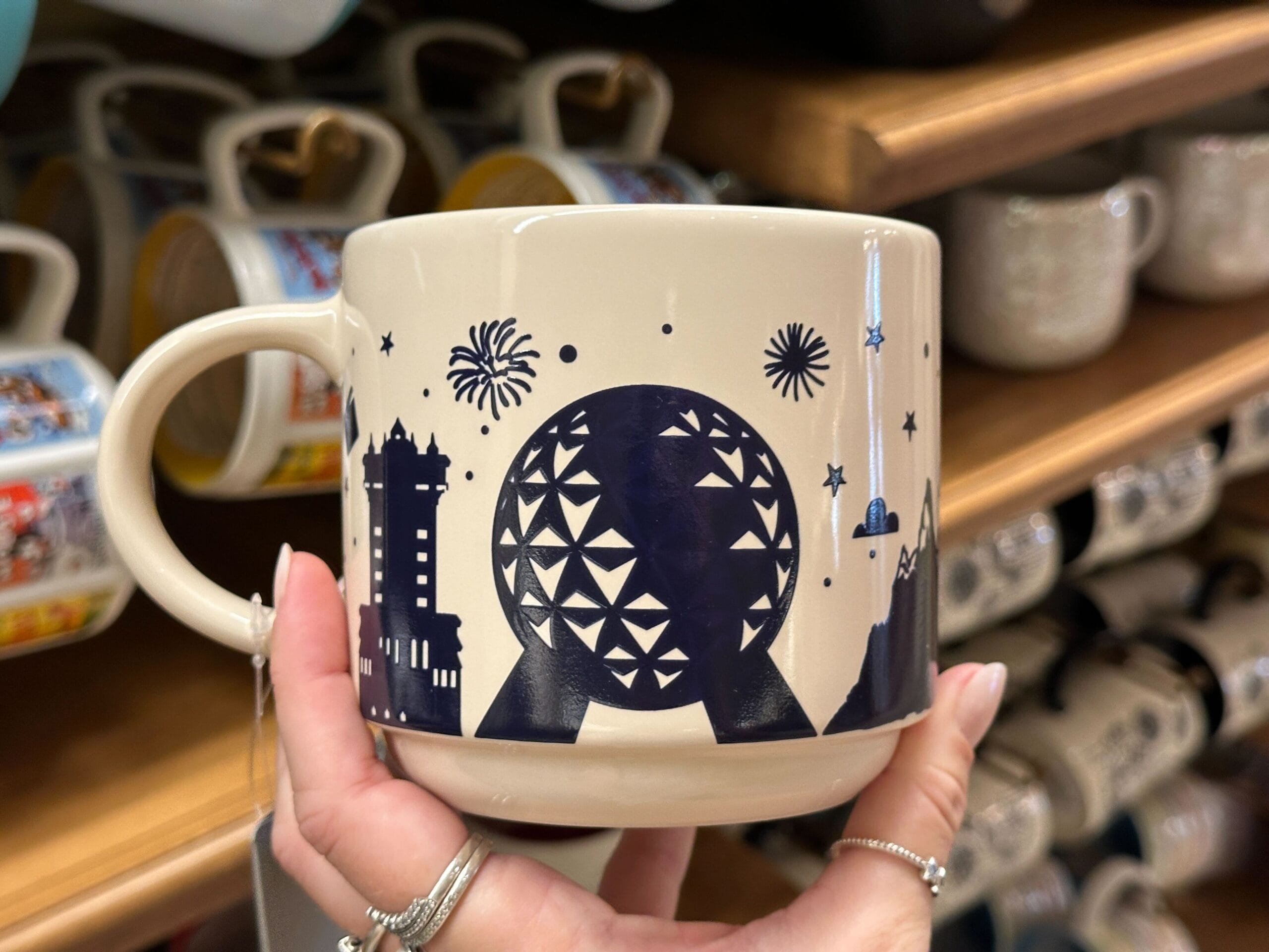 Hand holding a mug with dark blue Epcot, Minnie, and fireworks design on a cream background, displayed in a store.