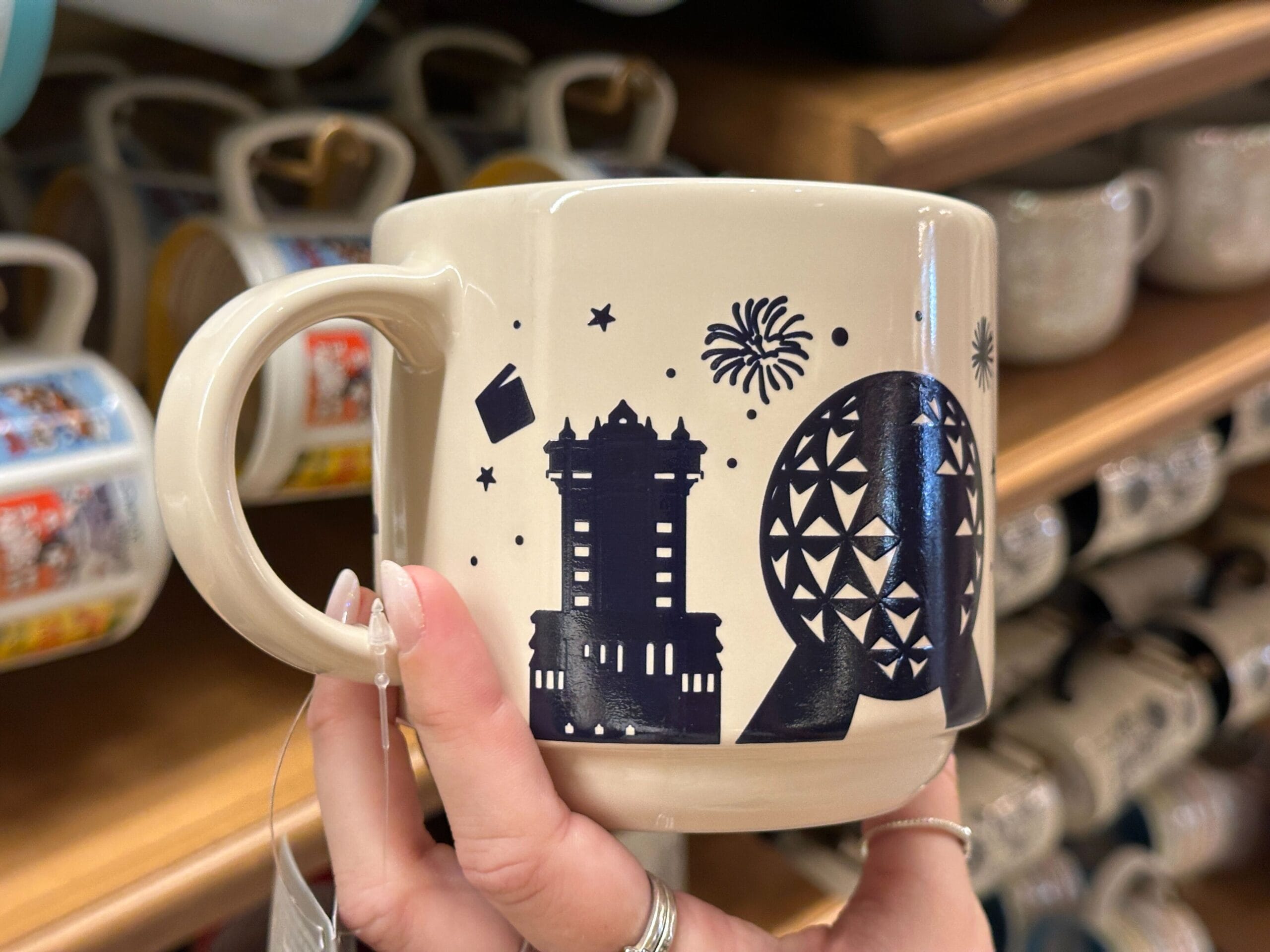 A hand holds a mug decorated with EPCOT’s Spaceship Earth, fireworks, and a tower featuring Mickey silhouettes.