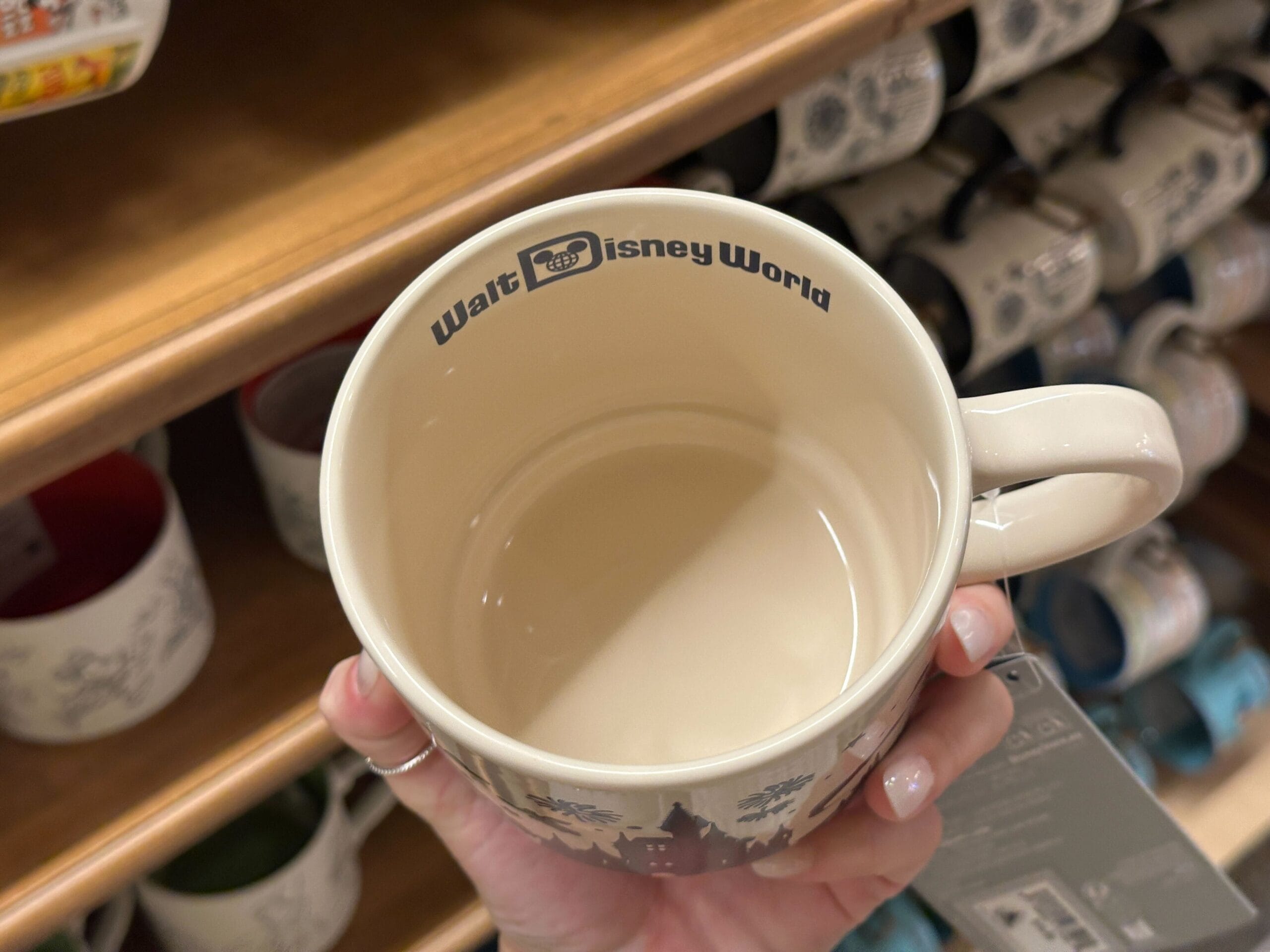 A hand holds a mug with “Walt Disney World” printed inside the rim, surrounded by Mickey mugs on the shelves behind.